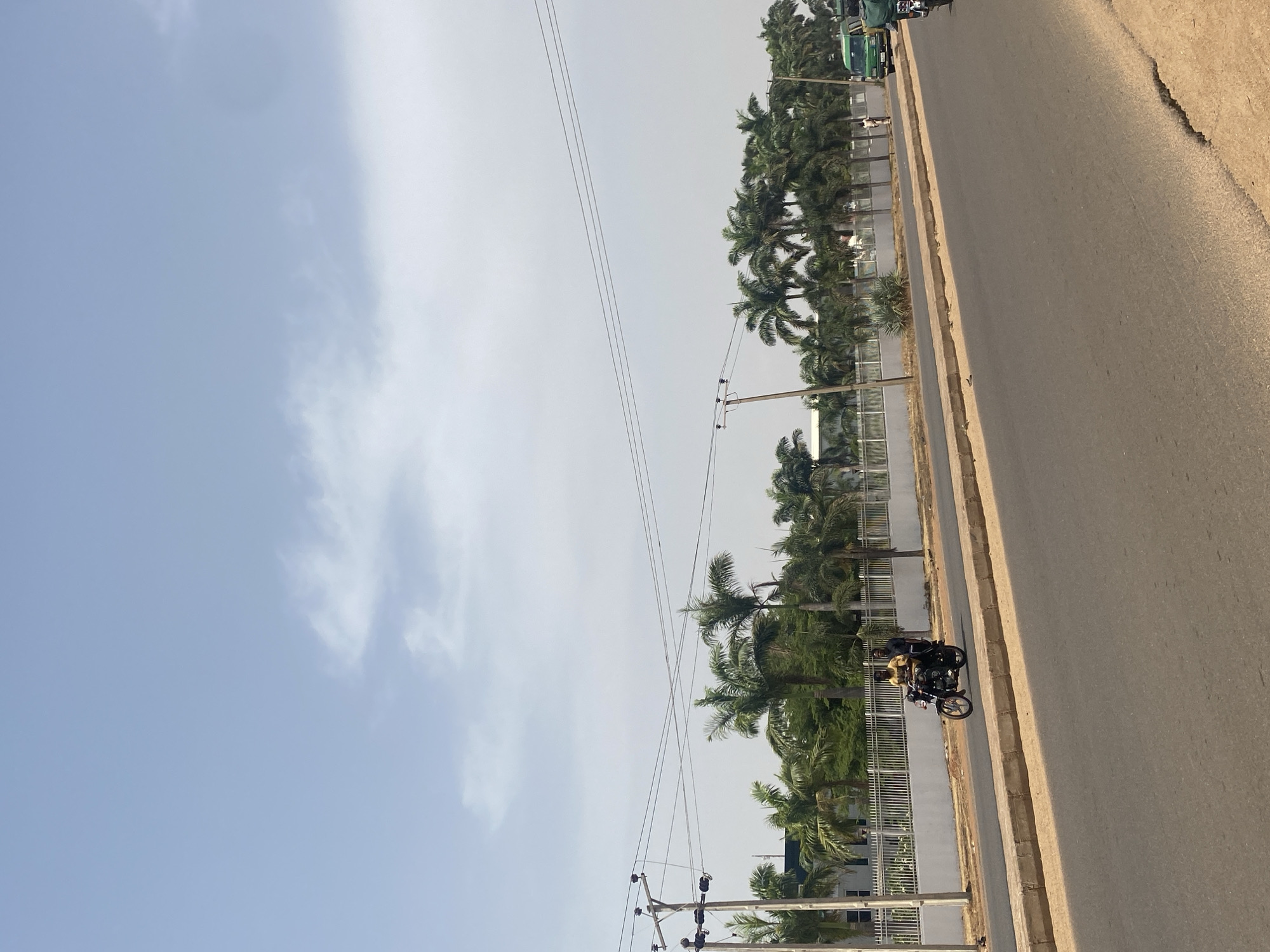 The image depicts a sunlit roadside scene in Rido, Nigeria. A two-lane asphalt road with a concrete median strip and dirt shoulders dominates the lower half of the frame. A dark-colored motorcycle, carrying two male occupants, is visible in motion on the left lane of the road. The driver wears a yellow shirt, and the passenger wears a dark shirt. Further down the road on the right, a green passenger van or bus is partially visible, with a solitary individual walking on the far dirt shoulder nearby.
Behind the road, a white perimeter fence extends horizontally across the scene, bordered by numerous tall palm trees and other dense green foliage. A large, multi-story building with dark roof elements is partially discernible behind the fence and trees. Multiple utility poles with intersecting overhead power lines are positioned along the left side of the road, extending into the background. The sky above is light blue with scattered white clouds.