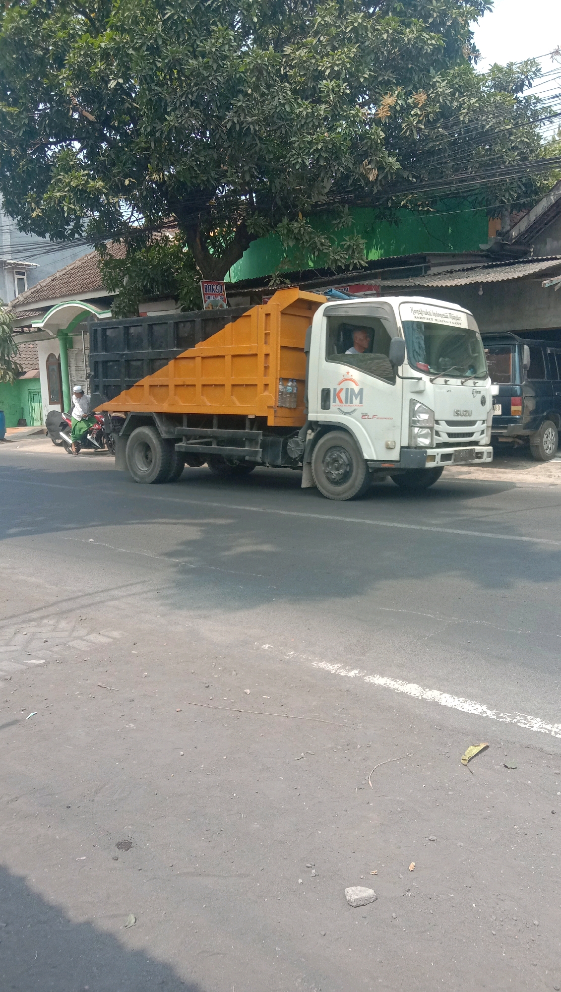 A white Isuzu Elf dump truck with an orange bed is traveling on a paved road in Karang Anyar, Indonesia. The truck has the logo "KIM" on its side. A man is visible in the driver's seat. In the background, there is a tree with green foliage, a building with a tiled roof, and a green wall. A motorcycle with a rider wearing a white cap and green clothing is also on the road in front of the truck. The road surface shows some debris and a white line marking. The scene appears to be in an outdoor urban or semi-urban environment.