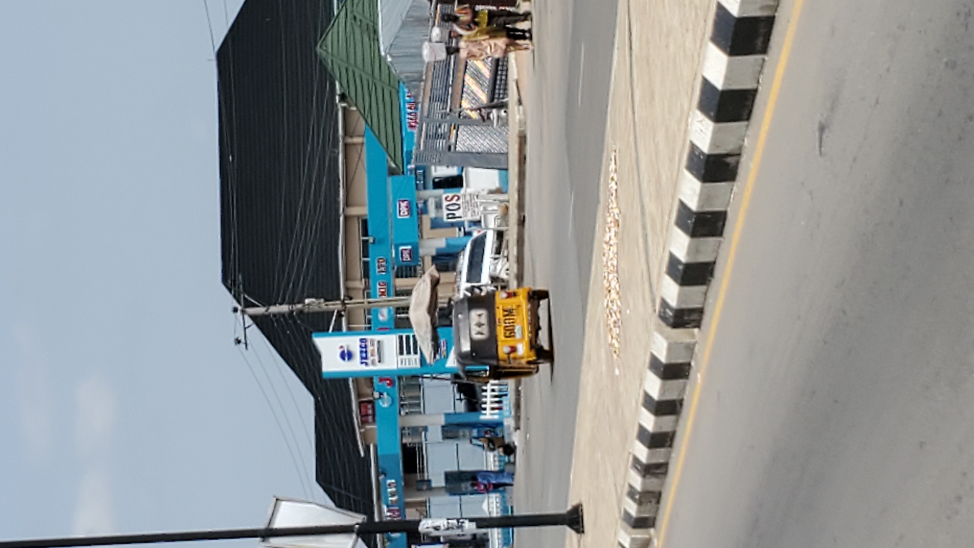 The image depicts a street scene in Jalingo, Nigeria, focusing on a Jezco fuel station. The fuel station, with its distinctive blue and white branding, features several pumps under a covered canopy. A yellow and black auto-rickshaw, with the number "600M" displayed on its rear, is driving away from the station on the paved road. In the background, a dark, pitched roof building is visible. To the right of the fuel station, a group of individuals, including women carrying items on their heads, are walking. The street is lined with a concrete barrier painted with alternating black and white stripes. The overall environment suggests an active commercial area with ongoing vehicular and pedestrian traffic.