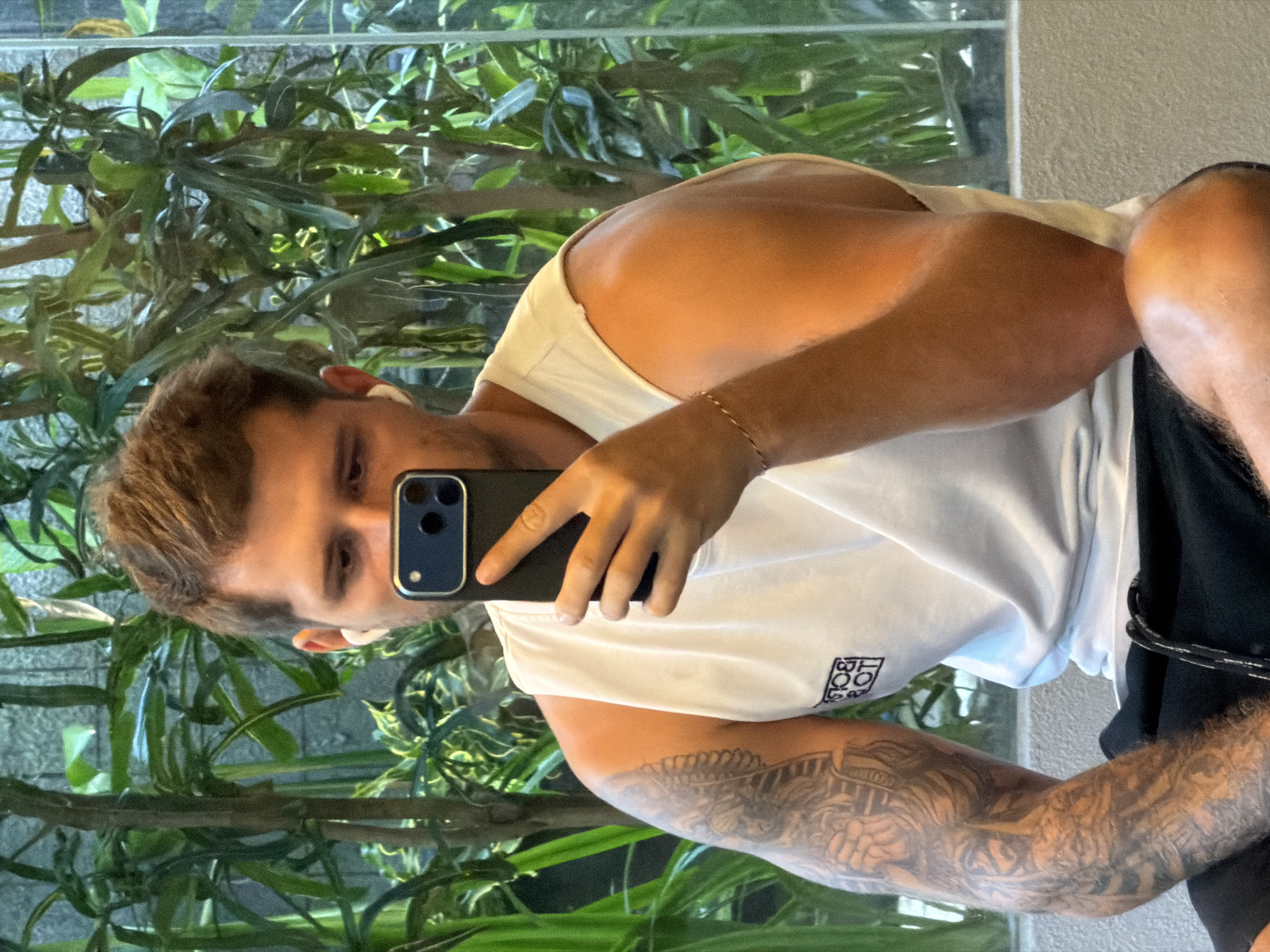 A light-skinned male takes a selfie in front of a mirror. He is wearing a white tank top and black shorts. He has a tattoo on his left arm. The reflection in the mirror shows plants and a wall. The location appears to be indoors in Kuta, Indonesia.