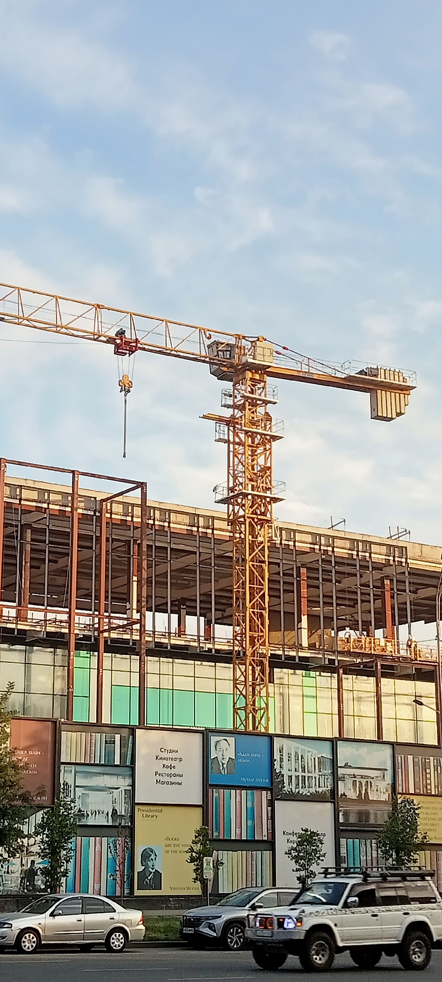 An outdoor, eye-level, medium shot captures a construction site in Almaty, Kazakhstan, under a partially cloudy sky. A large, yellow tower crane dominates the upper portion of the image, its boom extending horizontally across the frame. Below the crane, the skeletal structure of a multi-story building is visible, with exposed metal beams and unfinished floors. The lower facade of the building features a large graphic display designed to resemble bookshelves, with some panels depicting portraits and architectural renderings. In the foreground, a two-lane road is visible, with two cars driving on it. The left lane has a silver sedan, while the right lane has a white SUV with a roof rack. The overall scene depicts ongoing construction and urban development in Almaty.