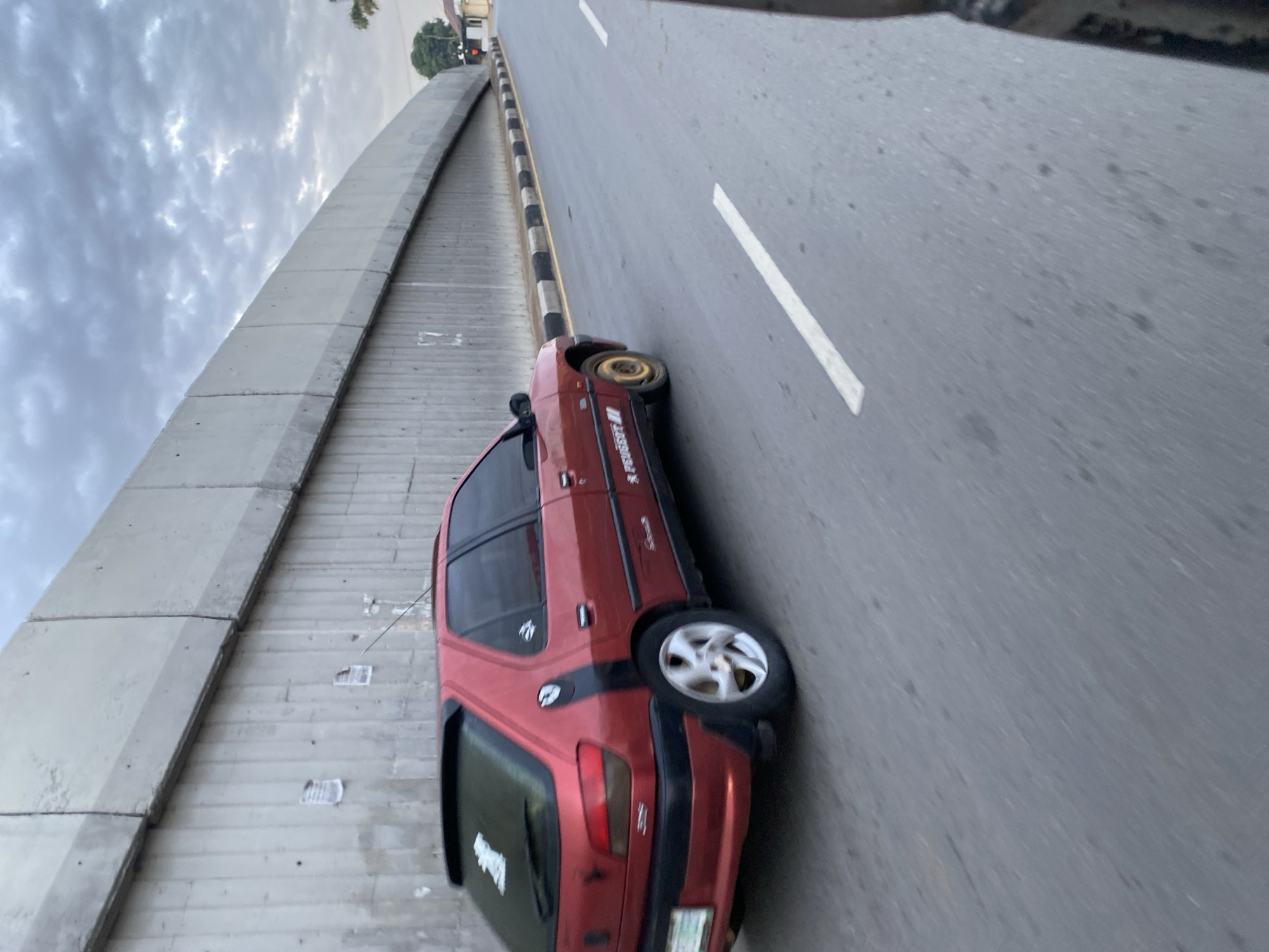 A red Peugeot 306 car is driving on a road in Jos, Nigeria. The car has a spoiler and stickers on the rear window and side. The road has white and black markings, and is bordered by a concrete barrier. The sky is cloudy. The scene appears to be a street view.
