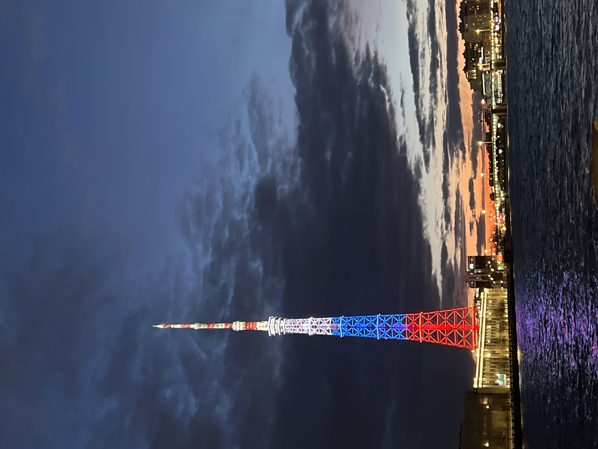 This image captures a nighttime or dusk scene in Saint Petersburg, Russia, centered around a strikingly illuminated television tower. The Saint Petersburg TV Tower, a tall, slender lattice structure, is brightly lit in the colors of the Russian flag: red at the base, white in the middle, and blue higher up, with the very top featuring more red and white segments.
The tower stands against a dramatic sky that transitions from a dark, deep blue at the top to lighter, warmer tones of orange and pink near the horizon, indicative of twilight. Streaks of dark clouds are visible across the sky.
To the right of the tower, a body of water, likely the Neva River or a canal, stretches across the foreground, reflecting the city lights in shimmering streaks of purple and white. Along the far bank of the water, a cityscape is visible, with numerous buildings glowing with warm artificial lights, suggesting an active urban environment. The buildings form a distinct horizon against the colorful sky.
No specific activities or people are discernible, and there is no visible text. The scene evokes a sense of grandeur and national pride, highlighting an iconic landmark of Saint Petersburg in a picturesque evening setting.