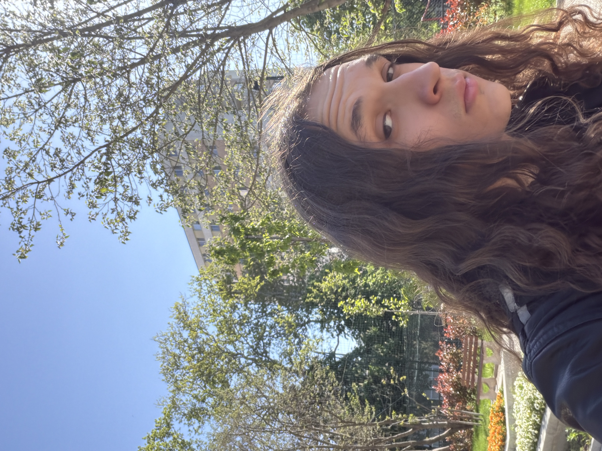 A selfie captures a young person with long, wavy brown hair outdoors in Plovdiv, Bulgaria, on a clear, sunny day. The person's face is seen from the forehead to below the chin, looking slightly upwards and to the right, with a neutral to contemplative expression, evidenced by visible forehead wrinkles. They are wearing a dark jacket or top. The background features a bright blue sky partially framed by numerous tree branches adorned with fresh, light green leaves, indicating spring or early summer. A tall building with many windows is visible in the upper background. To the person's right and behind them, a landscaped area is glimpsed, showing a wooden fence or railing and patches of red, orange, and white flowers amidst more green foliage, suggesting a park or garden setting within an urban environment. No text is visible in the image.