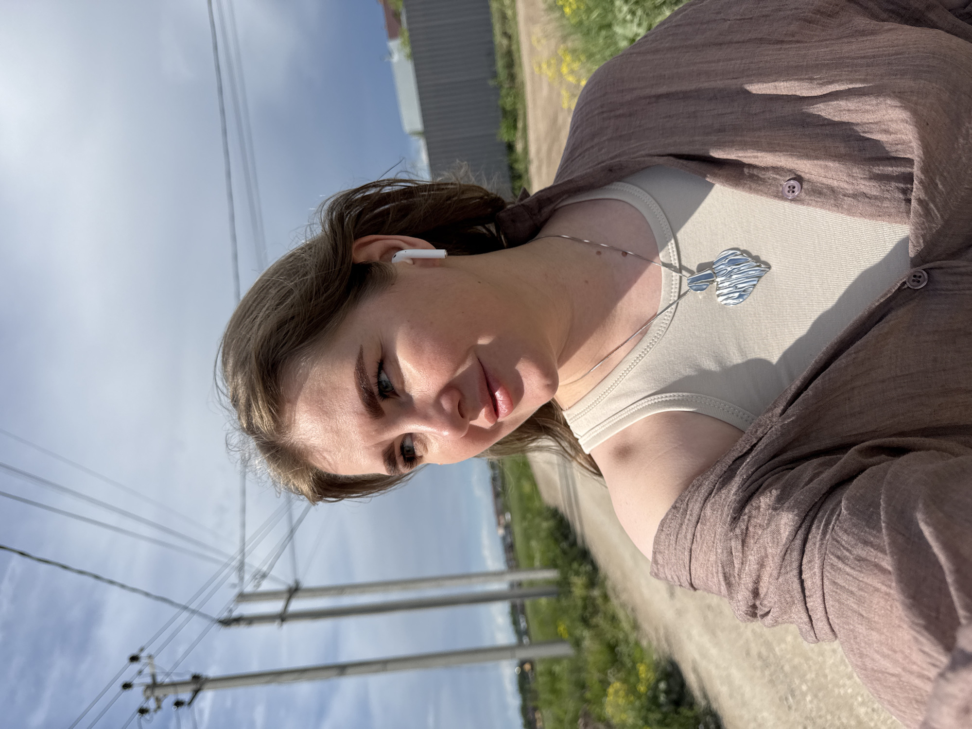 A selfie captures a woman outdoors in Горный, Russia. She wears a beige top, a brown shirt, and a silver necklace. An earbud is visible in her left ear. In the background, utility poles, a road, greenery, and a cloudy blue sky are visible.