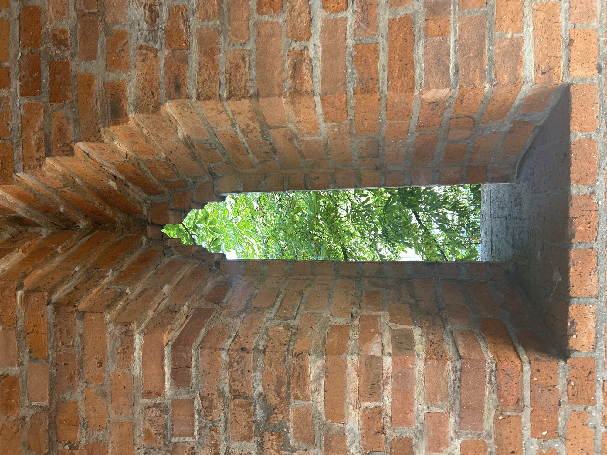 The image shows a narrow, tall slit window in a red brick wall. The window is shaped like an arch at the top and has a wider sill at the bottom. Through the window, lush green foliage from trees is visible against a bright sky. The brickwork is rustic and shows signs of age. This architectural feature is consistent with defensive structures found in historical buildings.
This particular type of window is a "shpil" window, a defensive architectural element found in the walls of the Moscow Kremlin. These narrow, vertically elongated windows allowed archers to shoot arrows while offering a small target to the enemy. They are characteristic of the Old English style of fortification.