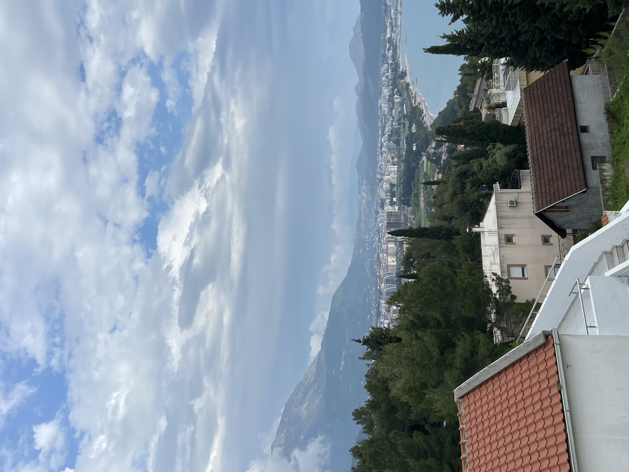 A panoramic view captures a coastal town or city in Šušanj, Montenegro, nestled between towering mountains and the sea under a partly cloudy sky. The image is a static landscape, showing a daytime scene, likely late morning or early afternoon, with bright but diffuse light.
The upper portion of the frame is dominated by a sky with scattered white and light grey clouds, some allowing patches of blue sky to show through. A prominent mountain range forms a dramatic backdrop, its slopes varying from verdant green to rocky outcrops, extending into the distance where they meet the water.
Along the coastline, a densely populated town or city stretches, with numerous buildings of various sizes visible. These structures appear to be predominantly residential, many featuring light-colored facades. The shimmering surface of the sea is visible to the right, bordering the urban development.
In the foreground, several buildings are distinctly visible, characterized by terracotta tiled roofs and light-toned walls. One particular building features a dark brown tiled roof and grey rendered walls. Tall, dark green trees, possibly conifers like cypress, are interspersed among the buildings and along the slopes, adding lush greenery to the landscape. External staircases and balconies can be seen on some of the foreground structures. No people or visible text are present in the image.