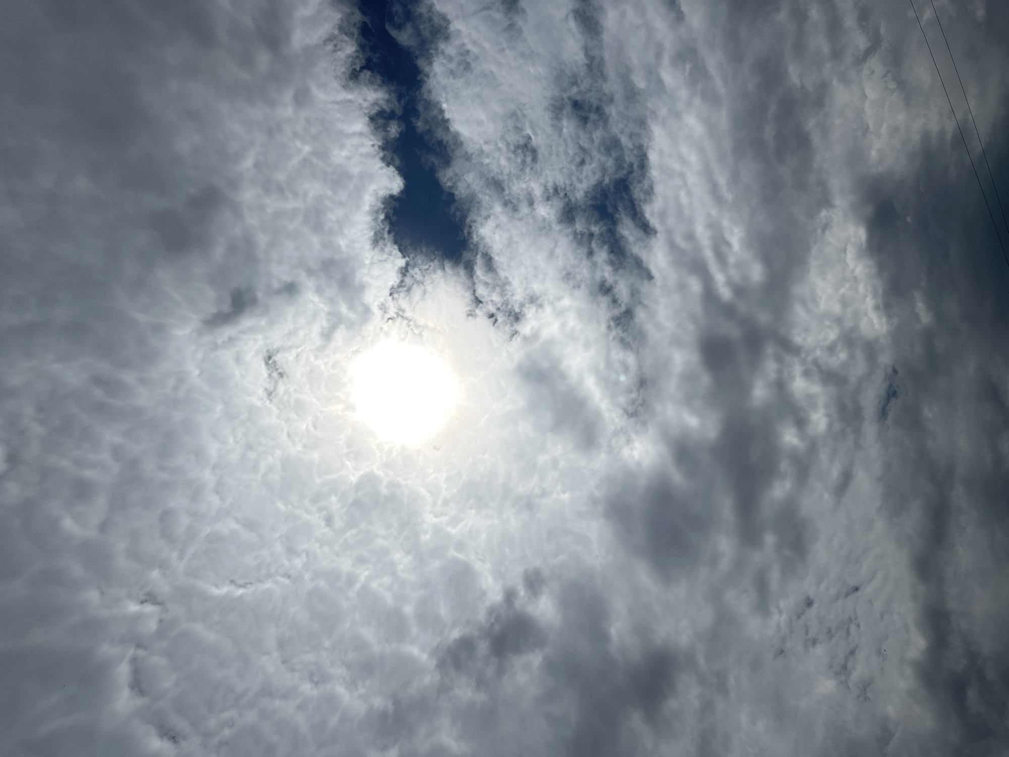 The image shows a sky filled with a variety of clouds, ranging from light and wispy to dark and dense. The sun is visible, shining brightly through a break in the clouds, creating a luminous effect. The overall impression is that of a dynamic sky, possibly before or after rain. There is no clear indication of the city of Târgu Neamț, Romania within the image itself, as it is a view of the sky only. The scene is devoid of any human presence, structures, or specific landmarks that would pinpoint a location within a city. The bottom right corner of the image shows two thin, dark, horizontal lines which are likely power lines or telephone wires, suggesting the presence of infrastructure.