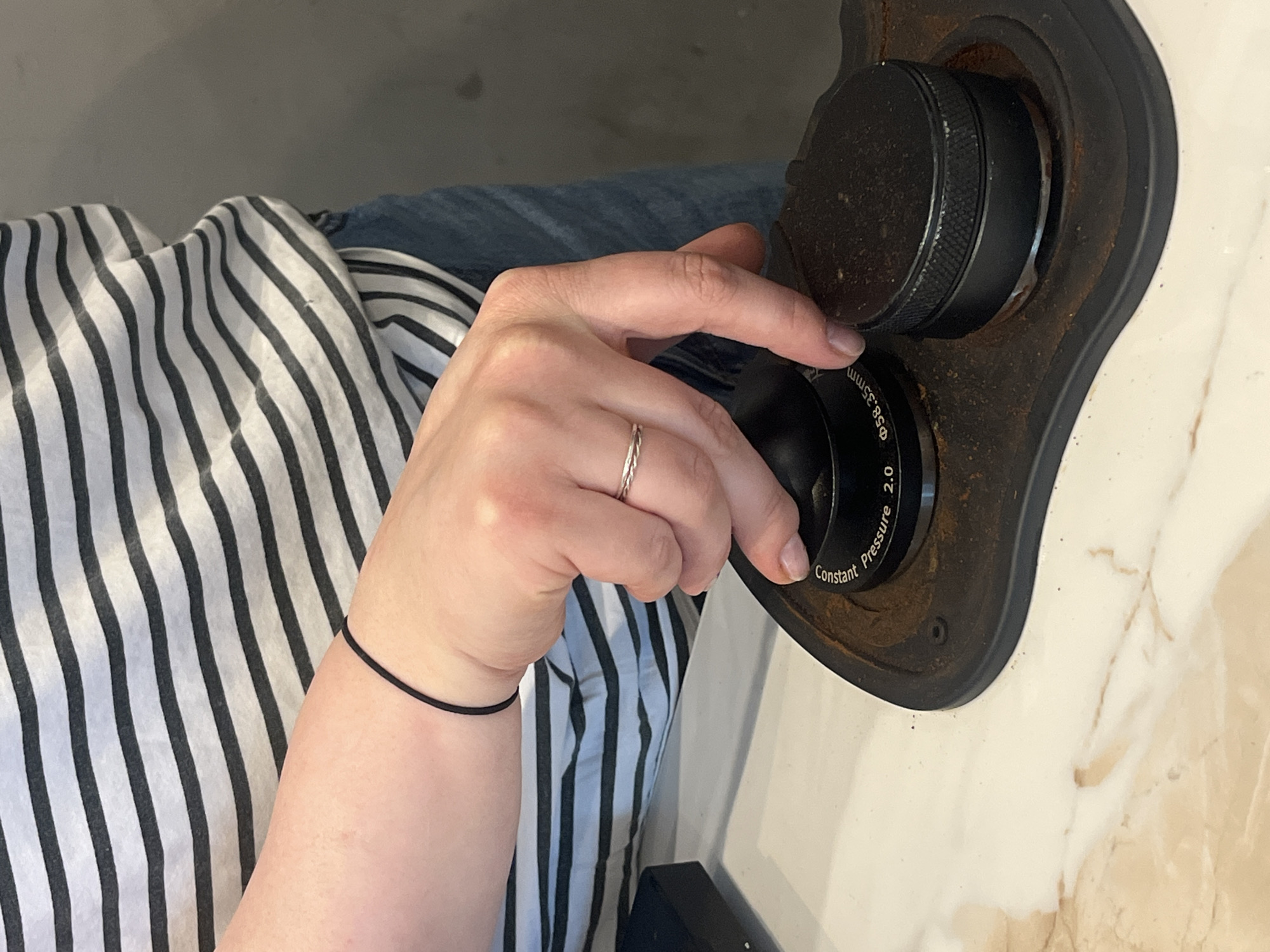A photograph depicts a person's arm and hand adjusting a lens. The person is wearing a striped shirt, blue jeans, a thin black wristband, and a silver ring on their ring finger. The hand is adjusting a black lens apparatus mounted on a rusty, black base. The lens has markings, including "Ø58.35mm" and "Constant Pressure 2.0." The background is a light, mottled surface, possibly a marble countertop. The location within the city of Budva, Montenegro cannot be determined.