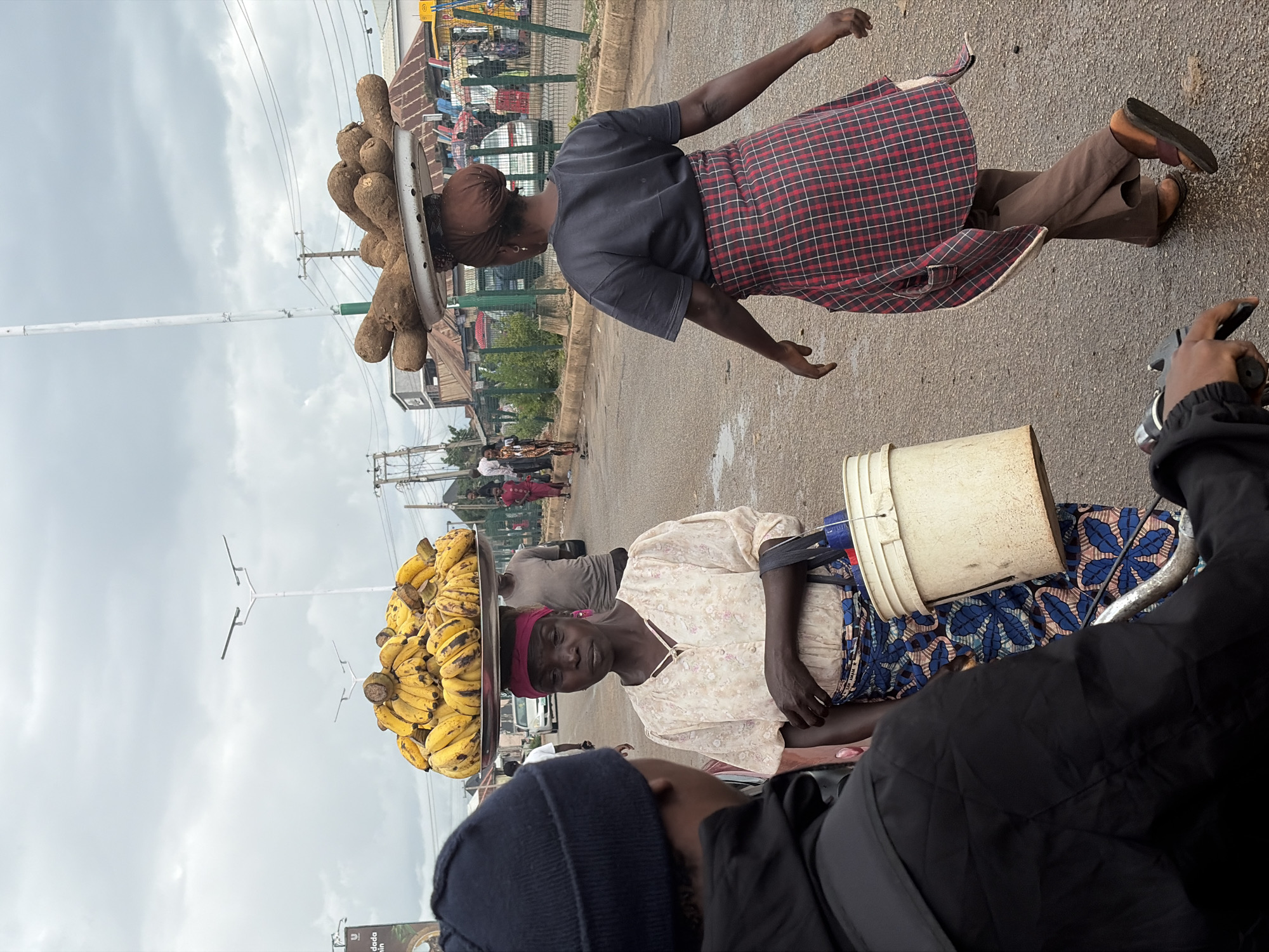 In Bukuru, Nigeria, a woman stands on a street carrying a large tray of bananas on her head. She wears a cream-colored blouse with a floral pattern, a blue and pink patterned wrap skirt, and a pink headscarf. She also holds a white bucket in her right hand. To her left, a person in a black jacket and navy blue beanie faces her. In the background, another woman walks away from the viewer, carrying a tray of root vegetables on her head. She wears a dark t-shirt and a red and black plaid wrap skirt. The street is paved and appears wet, suggesting recent rain. Buildings and a fence line the right side of the street. The sky is overcast.