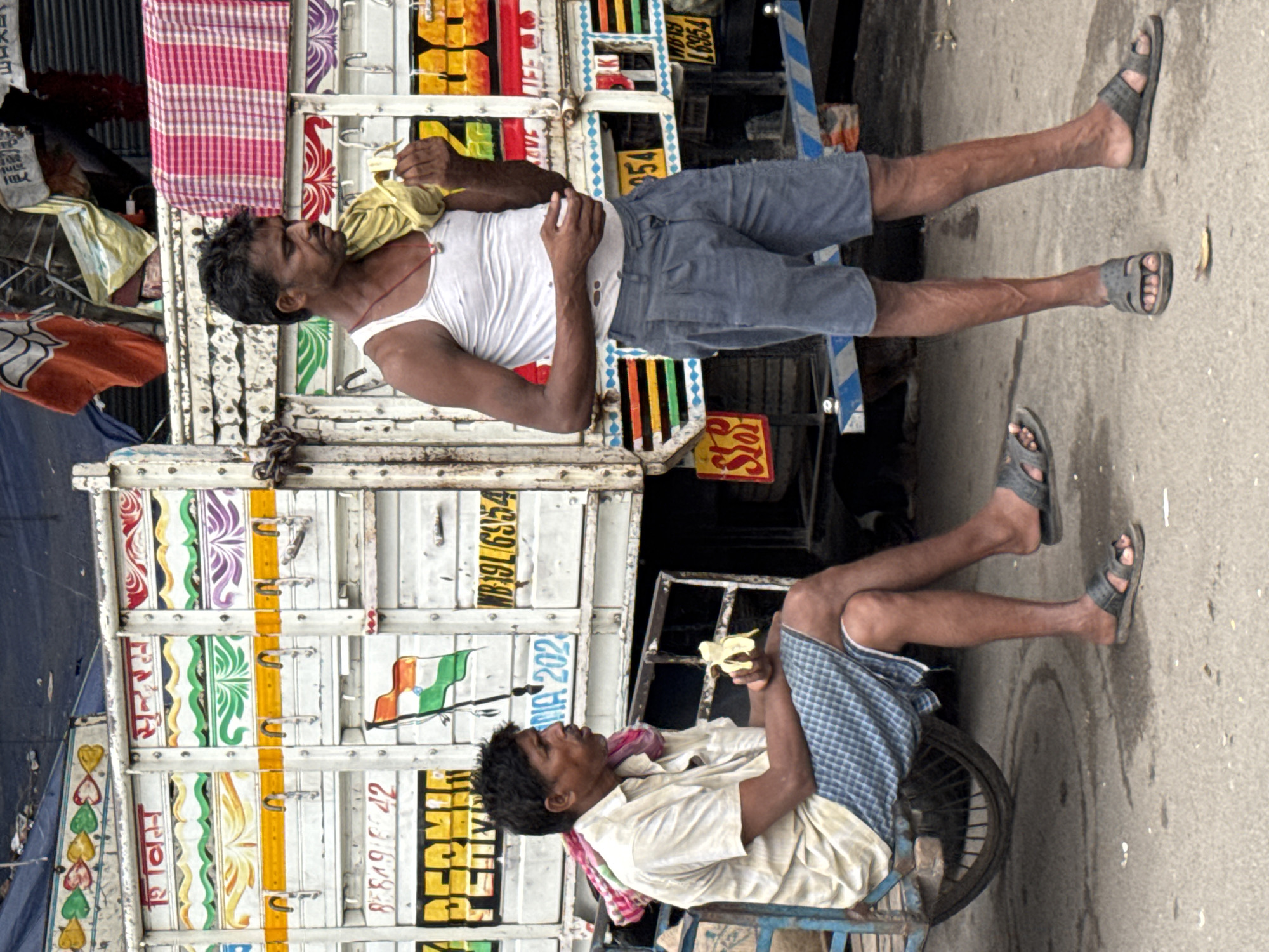In Kolkata, India, two men are near a brightly decorated truck. The man on the left, wearing a cream-colored shirt and blue checkered shorts, sits on a cart and eats a banana. A colorful cloth is draped around his neck. The man on the right, in a white tank top and grey shorts, stands holding a yellow cloth over his shoulder and a banana in his hand. The truck has various colorful paintings and writings on its sides, including what appears to be an Indian flag. The ground is asphalt, and the scene suggests a market or roadside setting in a bustling city environment.