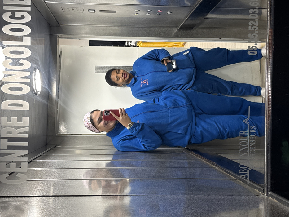 Two women in matching blue tracksuits pose in an elevator. The woman in the foreground holds a red smartphone, capturing the selfie. She wears a patterned head covering and a watch. The elevator reflects the scene, displaying text that reads "CENTRE D'ONCOLOGIE" and "ABONNOUR ASCENSEURS" along with a phone number. The elevator interior appears to be stainless steel with visible buttons and a glimpse of a yellow safety barrier.