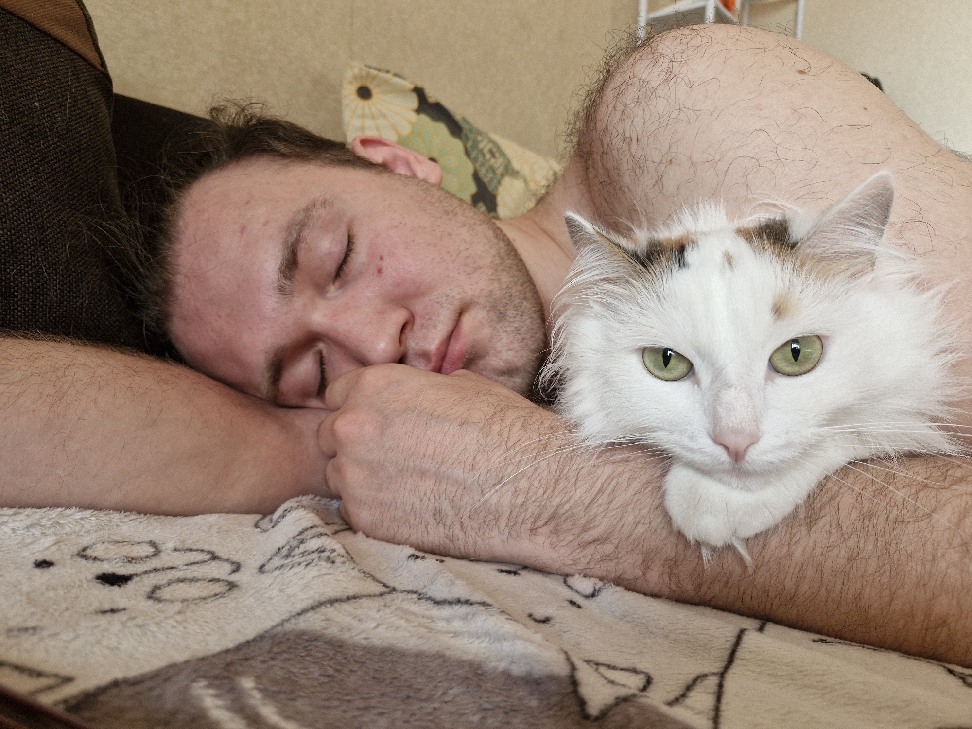 A man is asleep indoors, lying on his left side on a beige blanket with a cartoon cat design. His left arm is bent with his hand near his face. A white cat with patches of brown and black rests on the man's arm and chest. The location is likely in Yekaterinburg, Russia. The environment seems to be a domestic setting, suggesting a home. The man has short dark hair and a beard. The cat is looking directly at the viewer with its green eyes. A pillow is visible behind the man's head. The man's right shoulder is exposed, showing fair skin and hair.