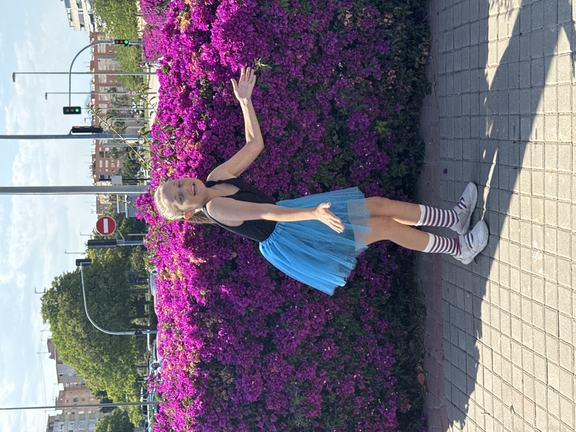 A girl stands in front of a hedge of purple flowers. She is wearing a black top and a blue tutu, along with striped socks and white sneakers. She has blonde hair and is smiling. Her right arm is raised in the air. The location appears to be an urban environment, as evidenced by the streetlights and buildings in the background. There is a brick sidewalk to the right.