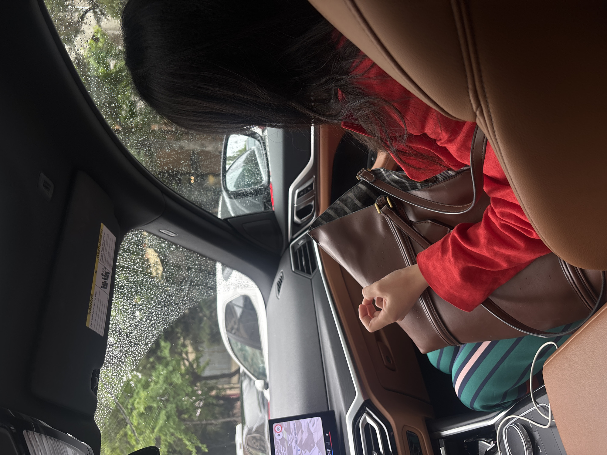 The image depicts a view from inside a car, presumably on a rainy day given the water droplets on the windshield. A person with long, dark hair and wearing a red long-sleeved shirt is reaching into a brown leather handbag. The person is wearing striped green and pink pants. A navigation system is displayed on a screen in the car's dashboard. Based on the GPS coordinates shown in the map display, the location appears to be in Yerevan, Armenia.