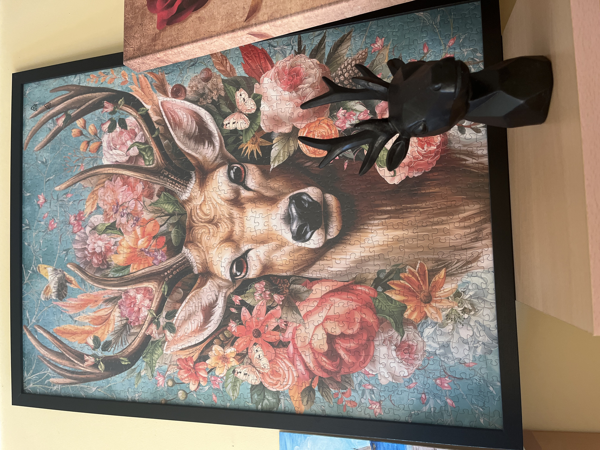 A close-up shot shows a framed jigsaw puzzle of a deer surrounded by flowers, hanging on a wall. A black, geometric deer head statue partially obscures the puzzle. The wall is painted a light yellow color. Other artwork is partially visible in the top left and bottom of the frame. The location is not discernible from the image.