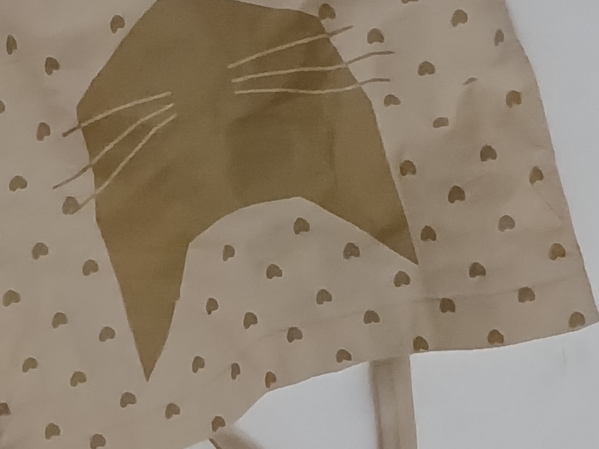 The image shows a close-up of a light beige fabric with a pattern of small, brown heart shapes. A larger, solid brown design of a stylized cat face with whiskers is also present on the fabric. The edges of the fabric are visible, and a portion of a light beige strap or ribbon is seen attached to it. The background appears to be a plain, light-colored wall. There is no information regarding a specific location in Buenos Aires, Argentina, or any people, actions, or interactions.