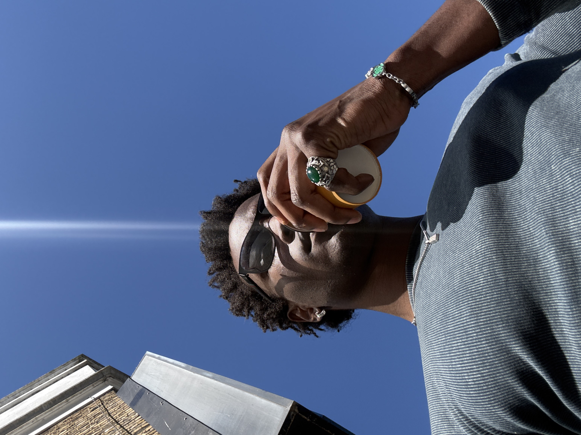 A low-angle, outdoor shot depicts a Black man holding a yellow cup. He is wearing sunglasses, a ring with a large green stone, a silver bracelet with green stones, and a gray and white striped shirt. He also has a silver chain around his neck. He is seen against a clear blue sky, with the corner of a building visible at the bottom of the frame. The location is potentially in London, United Kingdom.