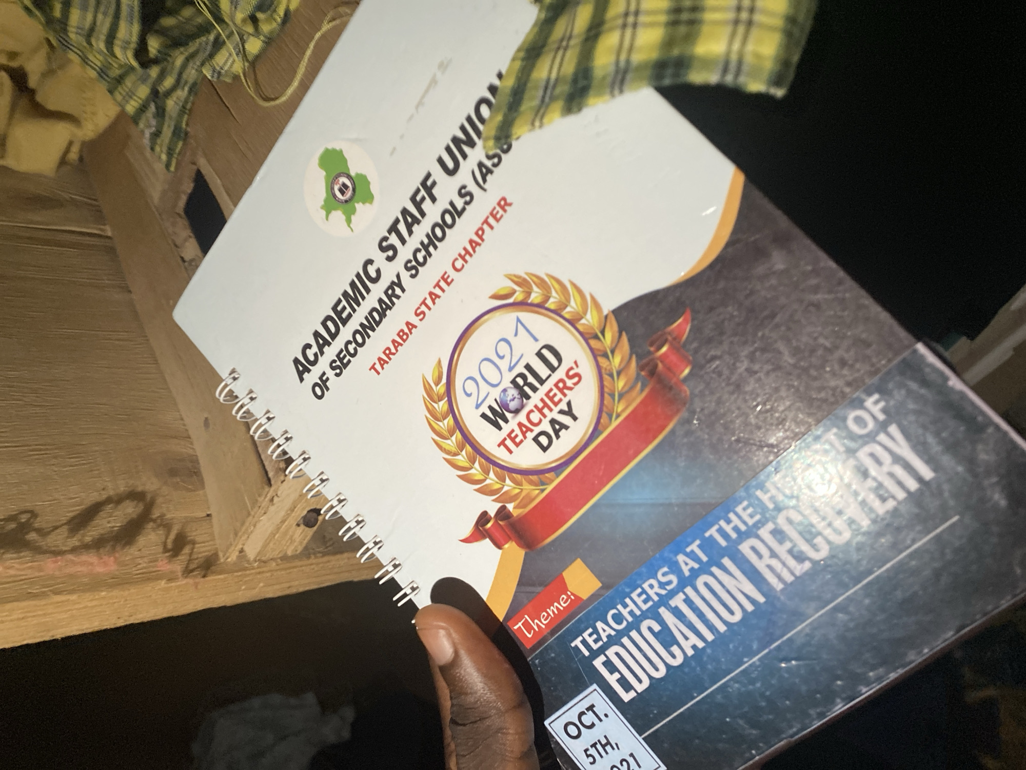 The image shows a close-up of a notebook titled "ACADEMIC STAFF UNION OF SECONDARY SCHOOLS (ASCS) TARABA STATE CHAPTER". The notebook is open, revealing a page with graphics related to "2021 WORLD TEACHERS' DAY". Below this, the theme for the event is stated as "EDUCATION RECOVERY", with "OCT. 5TH, 2021" also visible. A person's dark-skinned hand is holding the notebook. The notebook is placed on a wooden surface, possibly a table or shelf, with some out-of-focus fabric in the background. The lighting suggests it could be indoors, possibly at night or in a dimly lit area. The overall scene appears to be a casual presentation of the notebook.
