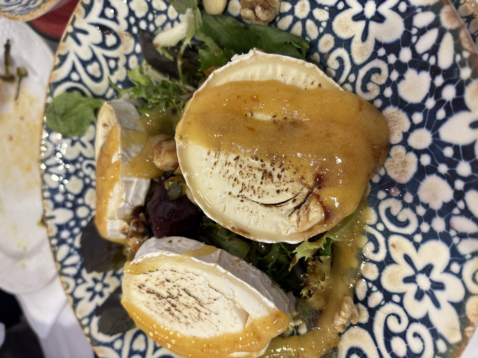 A close-up, overhead view depicts a prepared dish served on a patterned ceramic plate, set in a dining environment in Córdoba, Spain. The plate features a dark blue and off-white floral and scroll motif around its rim. The central subject is a salad composed primarily of three thick slices of goat cheese, each lightly browned on top and coated with a viscous, golden-orange glaze. Dark brown markings, possibly char or spices, are visible on the cheese surfaces. These cheese slices are arranged amidst a bed of mixed green leafy vegetables. Scattered throughout the salad are pieces of dark red beetroot, light brown walnuts, and smaller, lighter nuts, possibly hazelnuts or almonds, along with some green pistachio kernels. A generous amount of the golden-orange glaze or dressing pools around the ingredients on the plate. In the blurry background, parts of other white or off-white plates with food remnants are visible, suggesting a restaurant or dining table setting. No individuals are present in the frame.