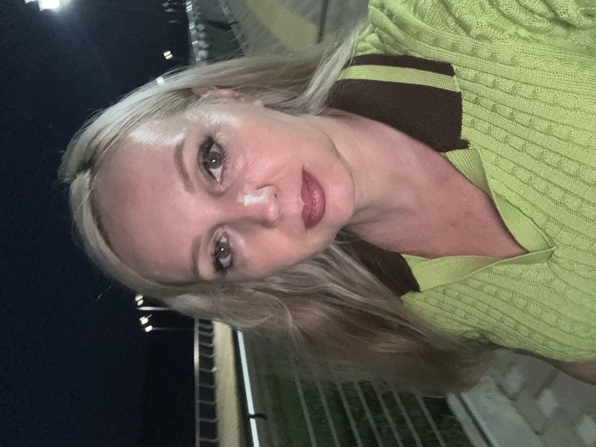 A blonde woman with light skin and dark red lipstick takes a selfie at night outdoors in Komsomolsk-on-Amur, Russia. She is wearing a light green knit shirt with a dark brown collar. The background shows a paved walkway with streetlights and some railing, indicating a public area.