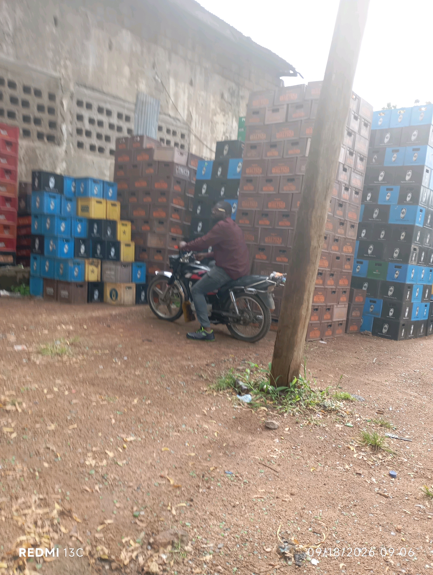 In Shendam, Nigeria, a man rides a motorcycle past stacks of beverage crates. The man, wearing a maroon long-sleeved shirt and gray trousers, is seated on a black motorcycle with a silver exhaust. He is positioned in front of tall stacks of brown and blue plastic crates, some of which are labeled with orange text. The crates are arranged in a haphazard manner, forming large walls. The ground is covered in reddish-brown dirt with scattered dry leaves and some green vegetation. Behind the crates, a building with a textured concrete wall and ventilation blocks is visible. The sky is overcast and bright. The image appears to be taken outdoors in what might be a storage or distribution area.