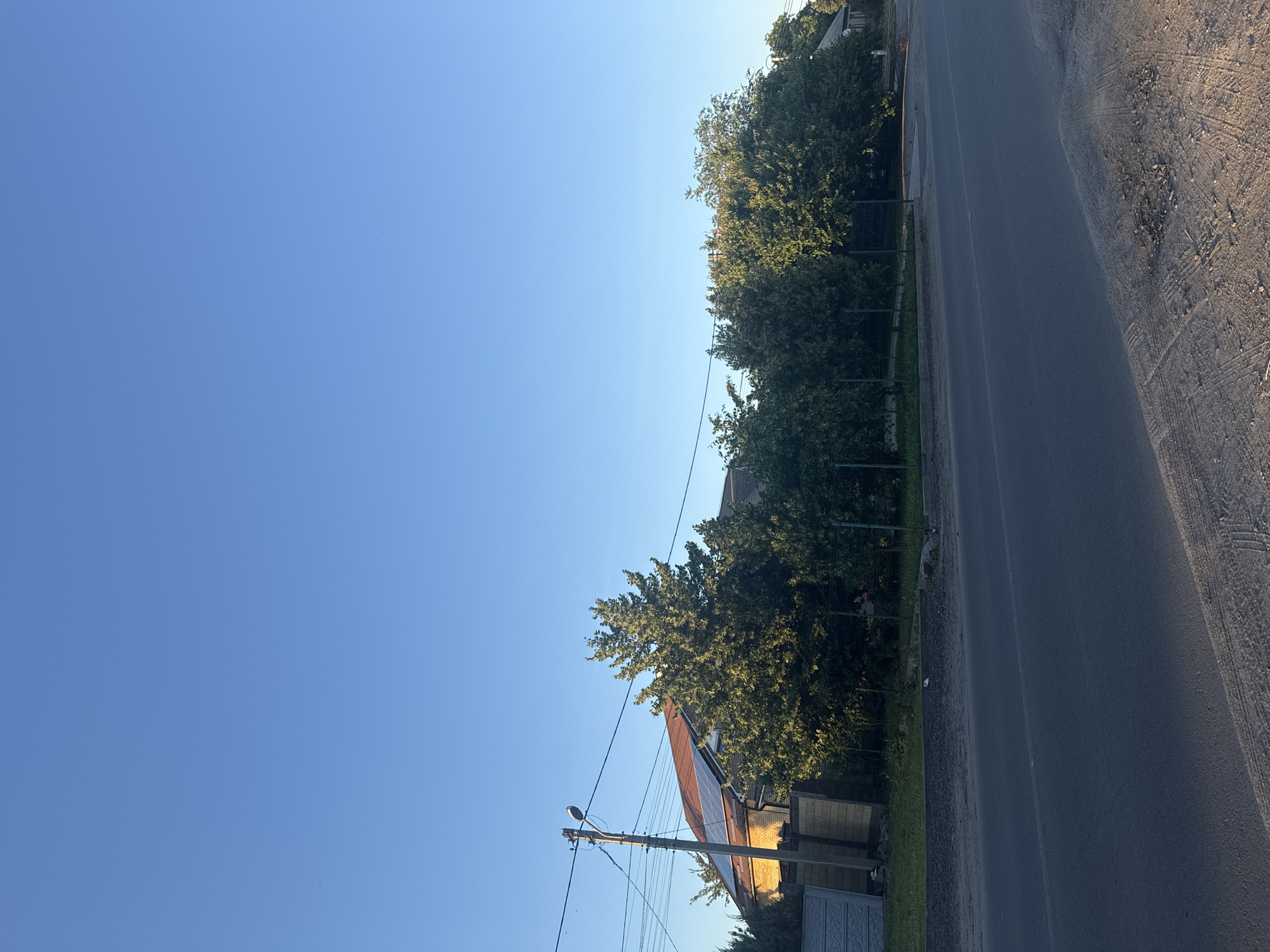 The image shows a street scene with a clear blue sky. On the left, there is a residential building with a brown roof and solar panels. A utility pole with wires and a street lamp stands in front of the building. A fence made of concrete blocks and metal mesh borders the property. Several trees line the street, with their foliage casting shadows. The road in the foreground appears to be paved asphalt, with some gravel and tire tracks on the shoulder. The overall impression is a quiet suburban or rural setting. The location is likely within the city of Dnipro, Ukraine, given the visual cues.