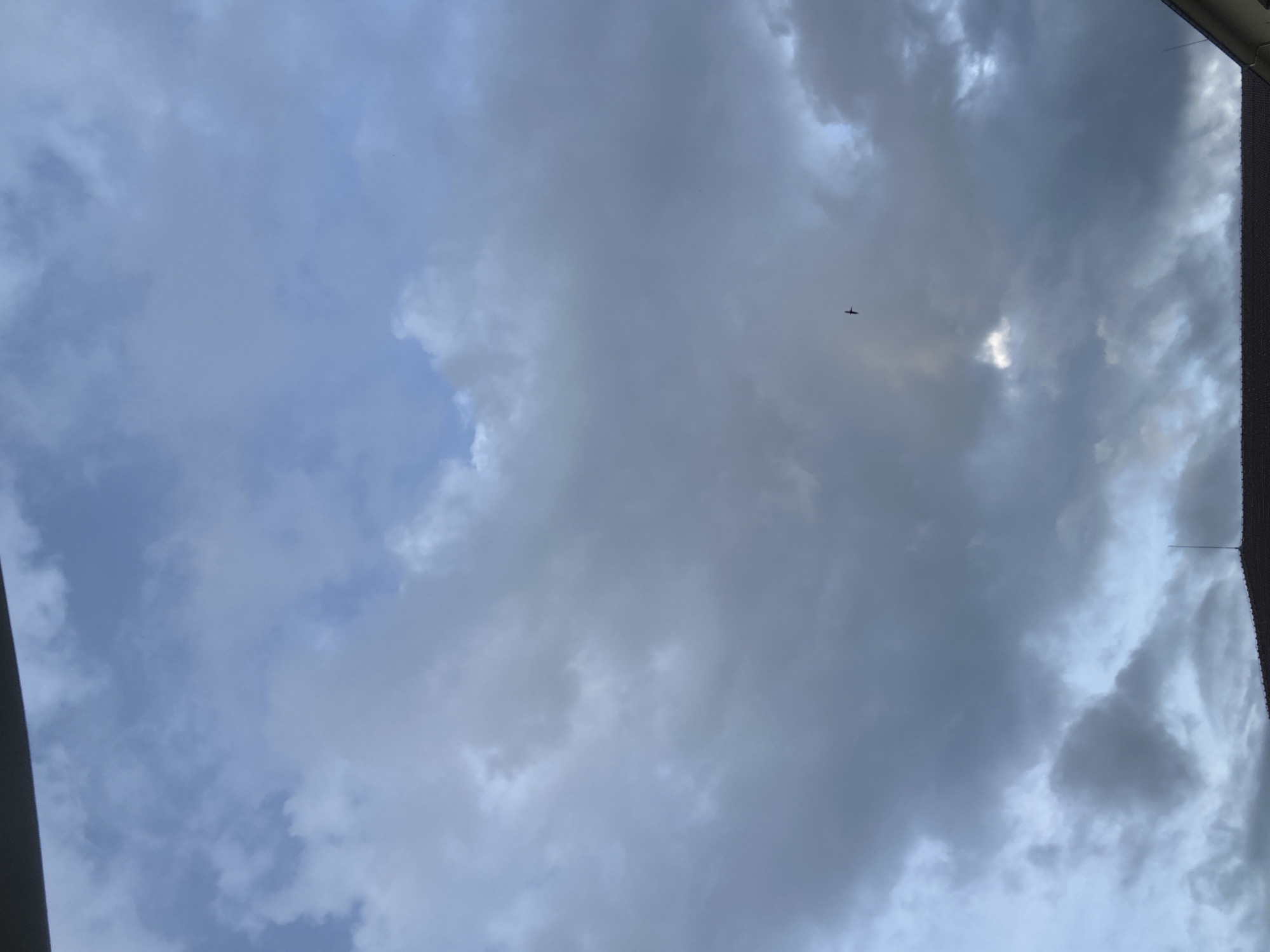 The image presents a sky view dominated by clouds, ranging from light grey to darker, denser formations. Patches of blue sky are visible, particularly towards the left and in some areas where sunlight appears to be breaking through the clouds. A small, dark silhouette of an airplane is visible in the upper-middle right section, flying horizontally across the frame.
The edges of the photograph show parts of a surrounding structure. On the far left, a dark, curved architectural element is partially visible. On the far right, a textured, dark vertical edge, likely a part of a building or window frame, extends into the shot. A thin, dark, vertical line, possibly an antenna or wire, can also be seen near the top right corner.
The scene depicts a cloudy daytime sky. The weather appears overcast or partly cloudy. No specific location cues for Troyan, Bulgaria, are visible within the image itself beyond the general sky, clouds, and airplane. There is no visible text.
