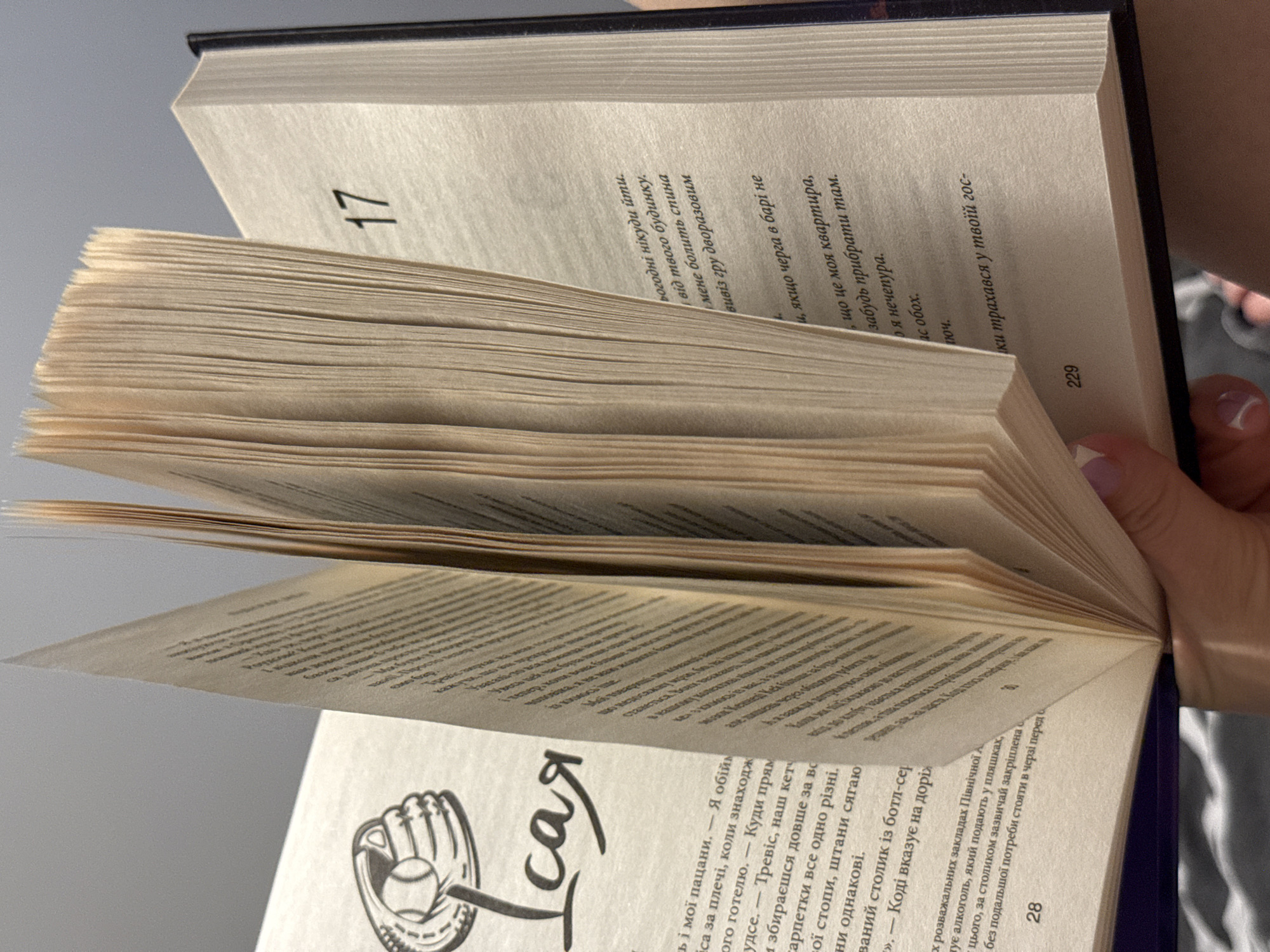 This media file shows a close-up, angled shot of an open book, held by a person's hand. The book has thick, slightly aged pages with black text printed in Ukrainian. The book is held open, revealing multiple pages stacked together, with a hand visible on the right side, gently holding the pages. The background is a plain, light gray.
Notable details include the visible page numbers "17" on one of the upper pages and "229" and "28" on lower pages. The text itself is in Ukrainian, and while specific phrases are visible, a full translation isn't possible from the image alone. Some of the text appears to be prose, and there are also some illustrations, including a drawing of a baseball glove and ball on one of the lower pages. The lighting suggests an indoor setting, with no indication of time of day or weather. The focus is entirely on the book and the act of reading or browsing its pages.