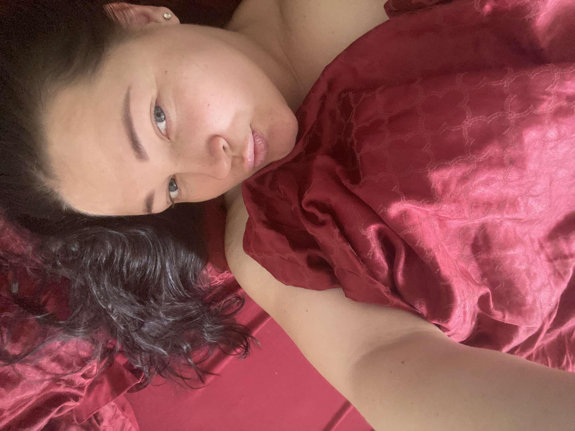 A woman is lying on a red surface, possibly a bed or similar soft surface. She is partially covered with a red, patterned fabric that appears to be satin or a similar silky material. The woman has dark hair and light skin. Her face is turned towards the camera. There is a single earring visible in her left ear. The background is primarily the red surface and fabric. There is no information in the image to determine a specific location, especially not within the city of Sochi, Russia.