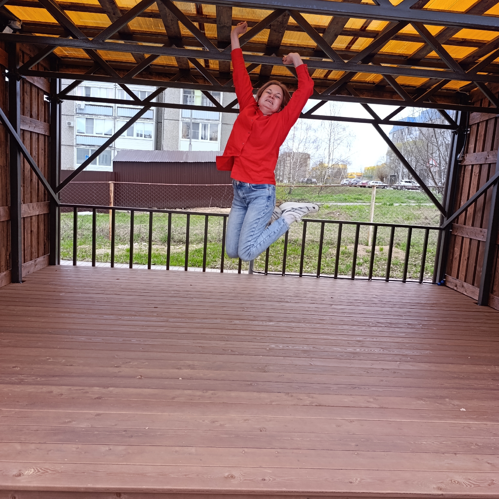 A dynamic outdoor shot captures a woman mid-air, jumping inside a wooden gazebo in what appears to be Марковский, Russia. The woman, dressed in a bright red long-sleeved shirt, light blue jeans, and white sneakers, has both arms raised above her head, her fingers slightly curled, as if reaching for or momentarily holding onto the yellow translucent roof structure of the gazebo. Her face shows a strained yet energetic expression.
The gazebo features a sturdy wooden floor made of planks laid horizontally, and dark metal support beams and railings that define its structure. The roof is a combination of dark wooden beams and a lighter, possibly corrugated plastic or plexiglass material, which casts a warm, golden hue. Beyond the railings, there's a patch of green grass, some bare trees, and a chain-link fence. In the background, several parked cars are visible on a road, and apartment buildings with multiple windows and balconies stand under a slightly overcast sky, indicating daytime. One building has visible air conditioning units. To the left, a dark brown shed or small building is also visible behind a fence. No discernible text is visible in the image.