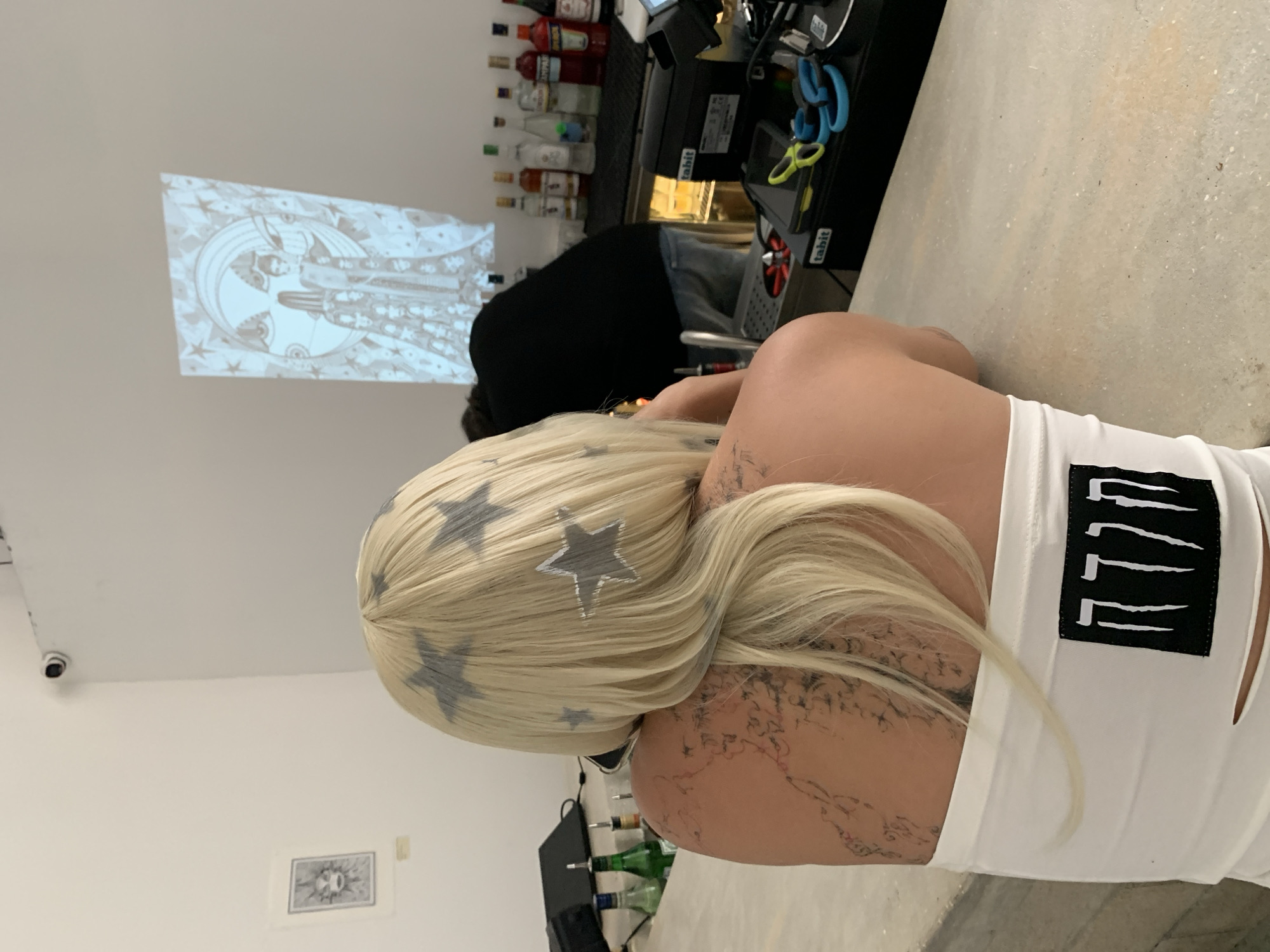 The image captures an indoor scene, likely a bar or event space in Tel-Aviv, Israel. The primary subject is a person viewed from behind, with long, blonde hair adorned with several grey star-shaped designs. They wear a white sleeveless top featuring a black patch on the back with a distinctive, stylized graphic composed of four vertical, claw-like marks. Extensive dark tattoos cover their right arm and shoulder. This individual is leaning on a light-colored, speckled counter or bar.
Behind the counter, another person, dressed in black, is partially visible, seemingly working in the bar area. The bar is stocked with numerous bottles of alcohol, including red, clear, and green varieties. Black bar equipment, some labeled "tabhit," and bright blue and yellow scissors are also present.
A monochromatic image is projected onto a white wall in the background, displaying a stylized moon or sun face surrounded by celestial patterns and stars. Below the projection, a smaller, framed black and white artwork, possibly an abstract eye design, is mounted on the wall. A small security camera is visible on the ceiling in the upper left corner of the frame. The artificial lighting suggests an evening or indoor event.