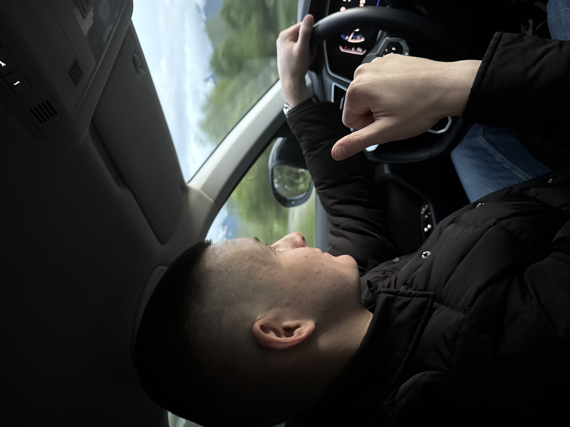 A man with a close-cropped haircut is shown in the driver's seat of a car. He is wearing a black puffer jacket and has one hand on the steering wheel. The other hand is extended towards the center of the frame, with the thumb and pinky finger extended in a "shaka" or "hang loose" gesture. The car is in motion, as evidenced by the blurred scenery visible through the windshield. The location is unknown.