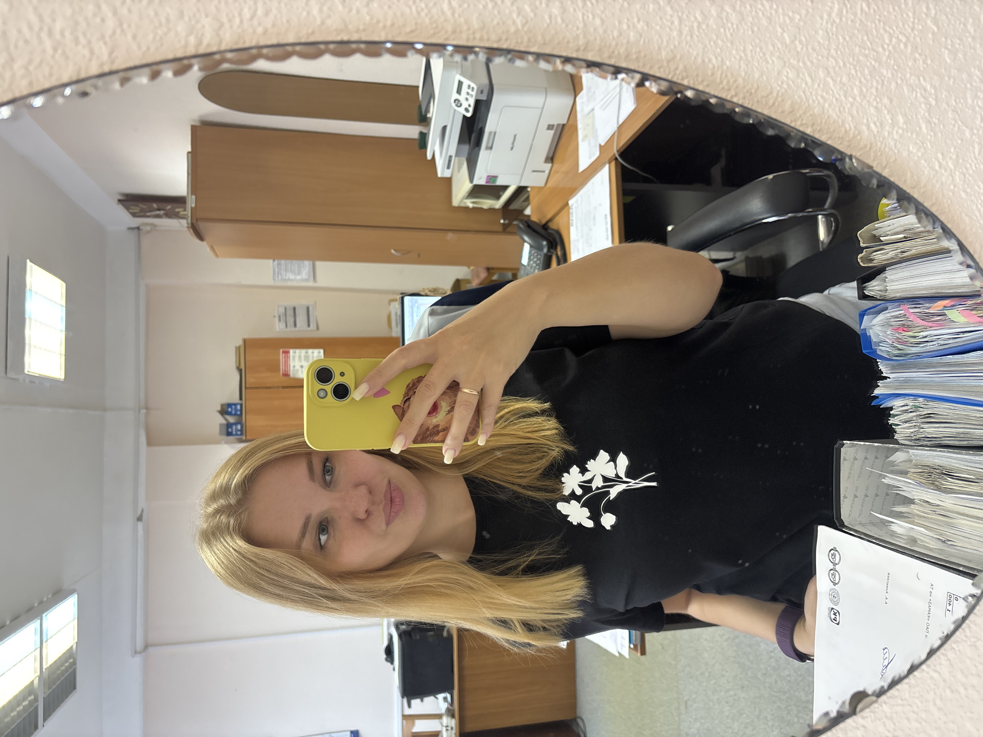 A young woman with blonde hair takes a mirror selfie in an office environment. She is wearing a black t-shirt with a white floral design. The office appears to be in Rusanovo, Russia, with beige walls, fluorescent lighting, and wooden furniture. In the background, there are filing cabinets, a printer, a computer monitor, and office supplies. The woman is holding a yellow smartphone to take the picture. Her nails are painted with a light color. A ring is visible on her right hand.
