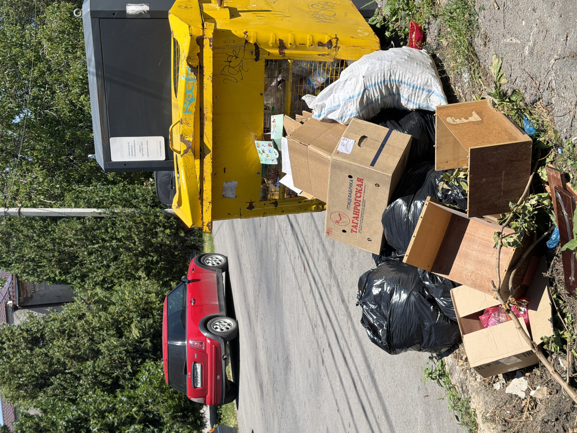 In an outdoor urban environment, likely on a street in Taganrog, Russia, a large yellow trash container, showing signs of rust and graffiti, is overflowing with garbage. Several black trash bags are piled beside it, along with cardboard boxes, one of which has text that includes "ТАГАНРОГСКАЯ". A white woven bag filled with trash also sits next to the container. In the background, a red Mini Cooper is parked on the asphalt road, with trees and a building visible behind it. The scene depicts an accumulation of refuse, suggesting a possible issue with waste management in the area. There are no people or animals visible in the image.