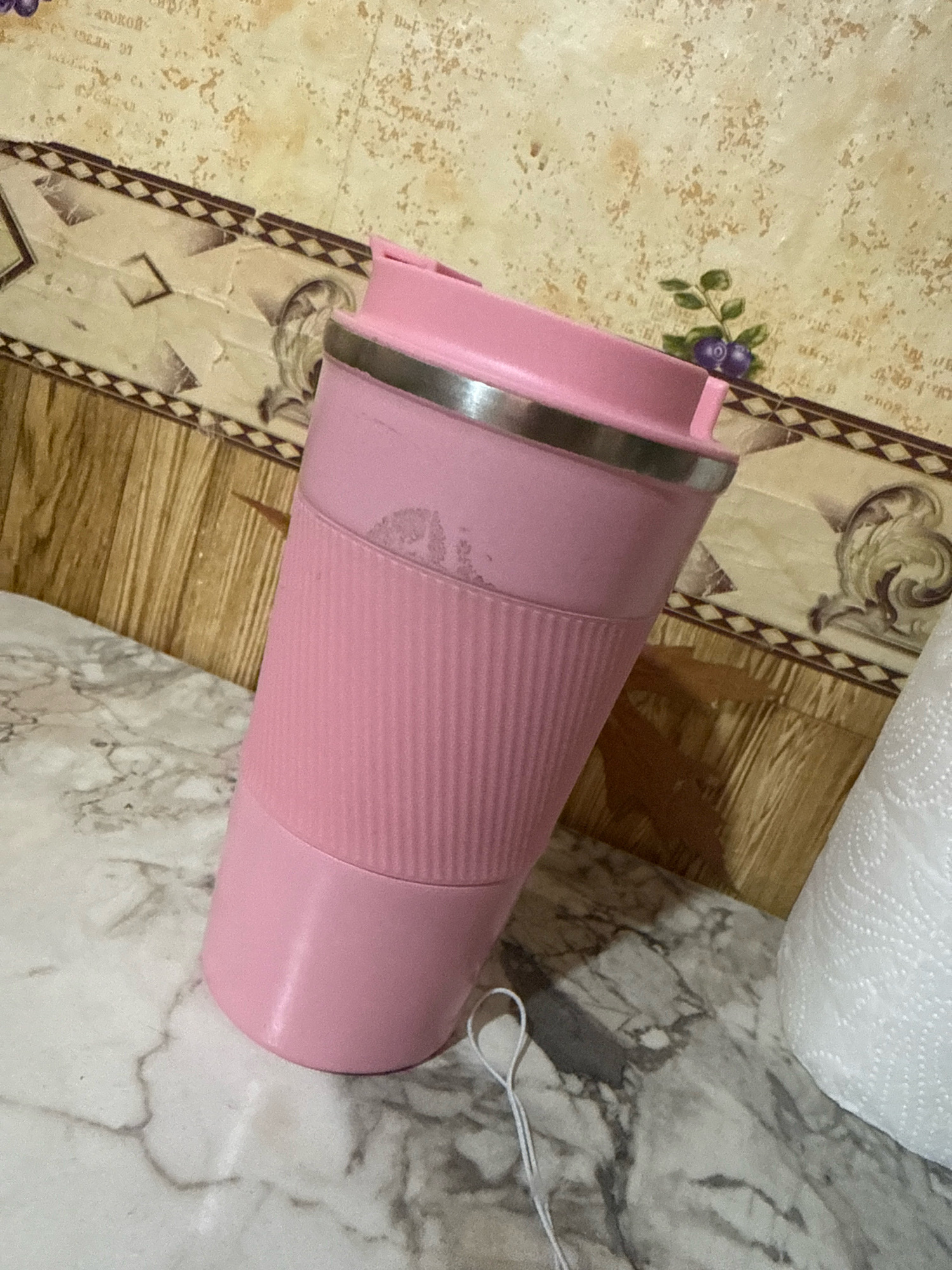 A pink tumbler with a silver rim and ribbed texture is positioned on a marble countertop. The background features a wall with a patterned wallpaper and wooden paneling. A roll of white paper towels is visible to the right of the tumbler. The scene appears to be indoors, possibly in a kitchen or dining area. There are no people or animals present.