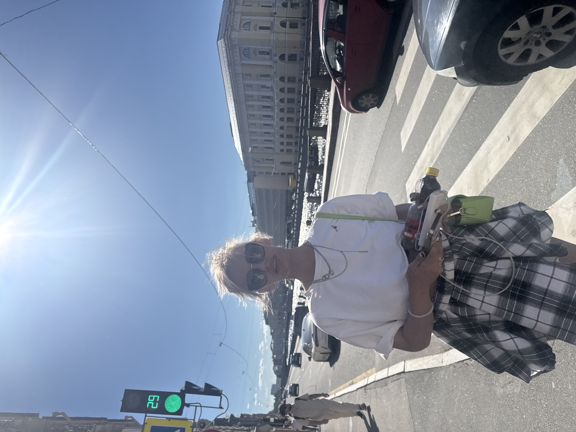 A woman stands on a sidewalk in Saint Petersburg, Russia, holding a bottle and other items. She is wearing a white t-shirt, a plaid skirt, and sunglasses. Behind her, a street with cars and a canal with a building on the other side is visible. A traffic light with a green signal and the number 29 is also visible. The sun is shining brightly, casting shadows on the ground.