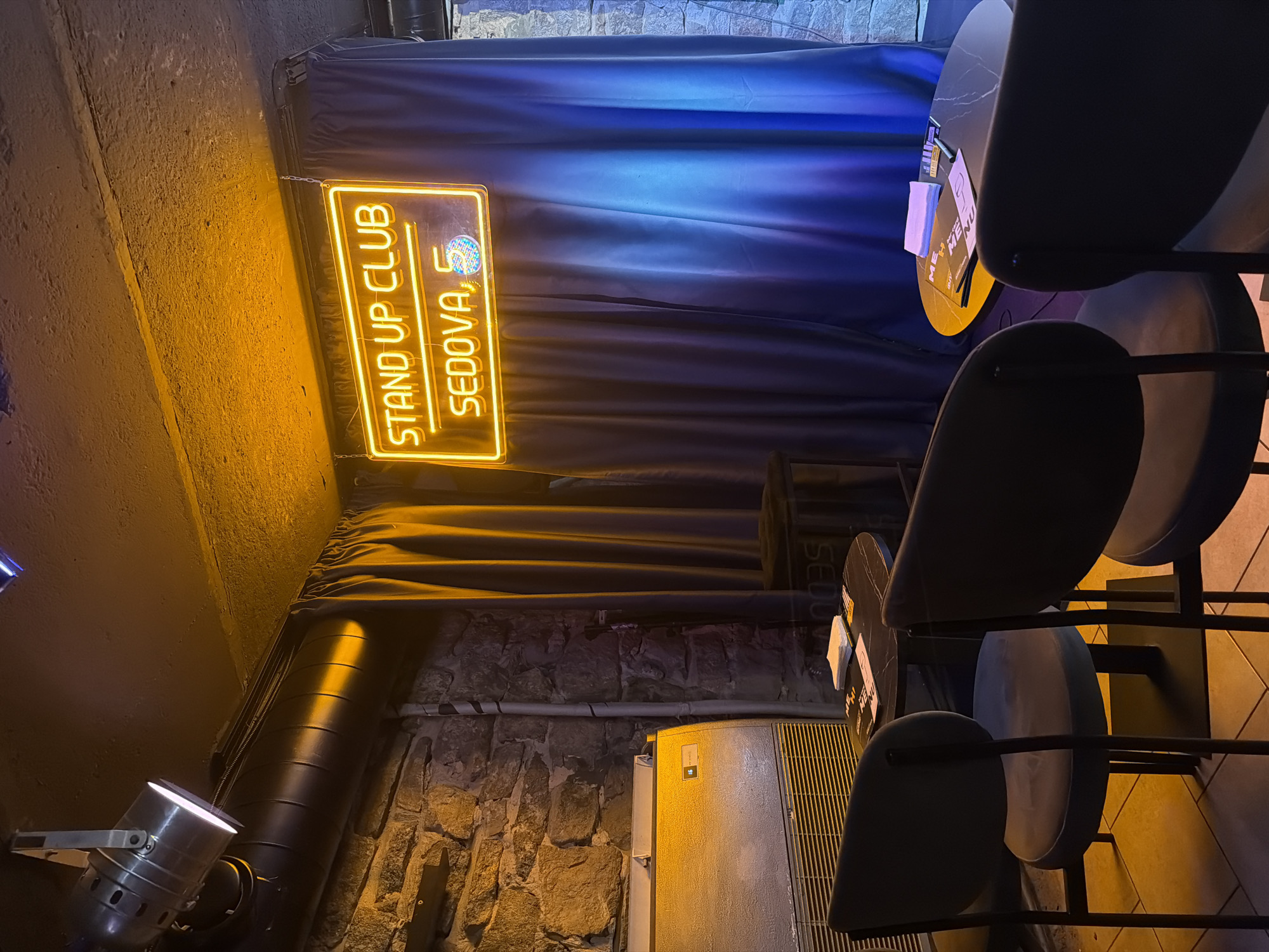 The image shows an indoor scene with a neon sign that reads "STAND UP CLUB SEDOVA, 5". The sign is illuminated in yellow and is hanging in front of a blue and purple gradient curtain. In the foreground, there are two dark-colored chairs and a round table with papers on it. To the left, there is a stone wall and what appears to be an air conditioning unit. A spotlight is visible on the ceiling, casting a warm yellow light on the scene. The overall atmosphere suggests a small, intimate venue, likely a comedy club or a similar entertainment space. The location of the "Sedova, 5" in Rostov-on-Don, Russia, further specifies the establishment.
