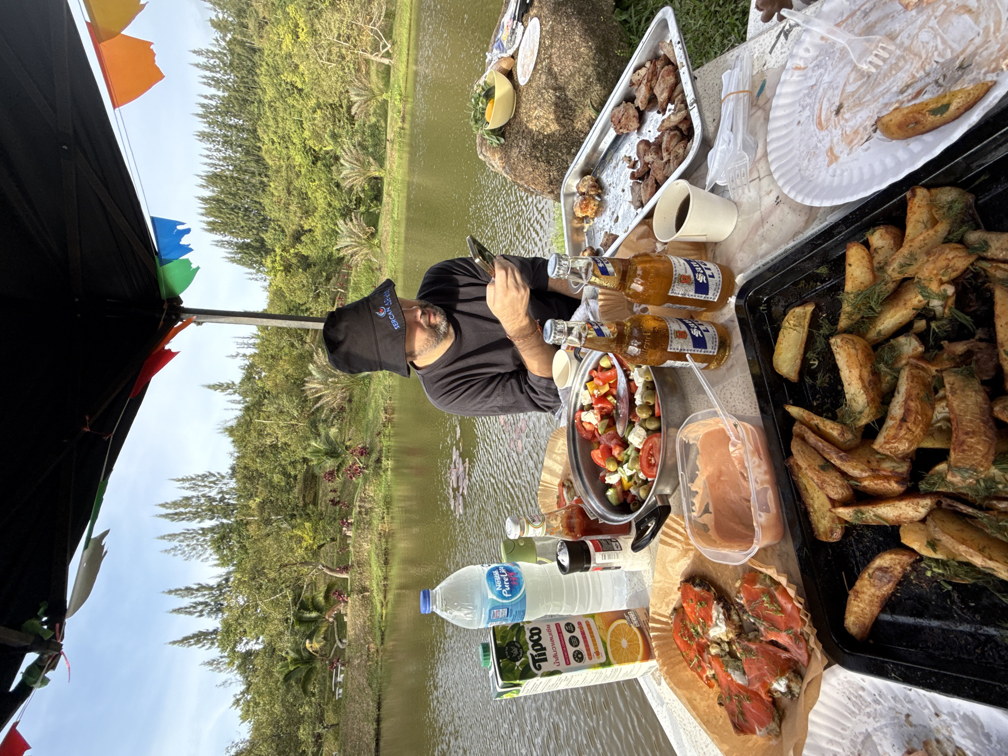 A man wearing a black bucket hat and a dark long-sleeved shirt sits at a table by a lake, looking at his phone. On the table, there is a meal laid out, including a salad, grilled meats, fries, and what appears to be smoked salmon on toast. There are also bottles of beer and a carton of orange juice. The setting appears to be outdoors, under a canopy, with trees and a hill in the background. The man's hat has a logo that reads "SERCAN AS". The location is identified as Ko Samui District, Thailand, indicated by the vegetation and architecture visible in the background, consistent with a tropical environment.