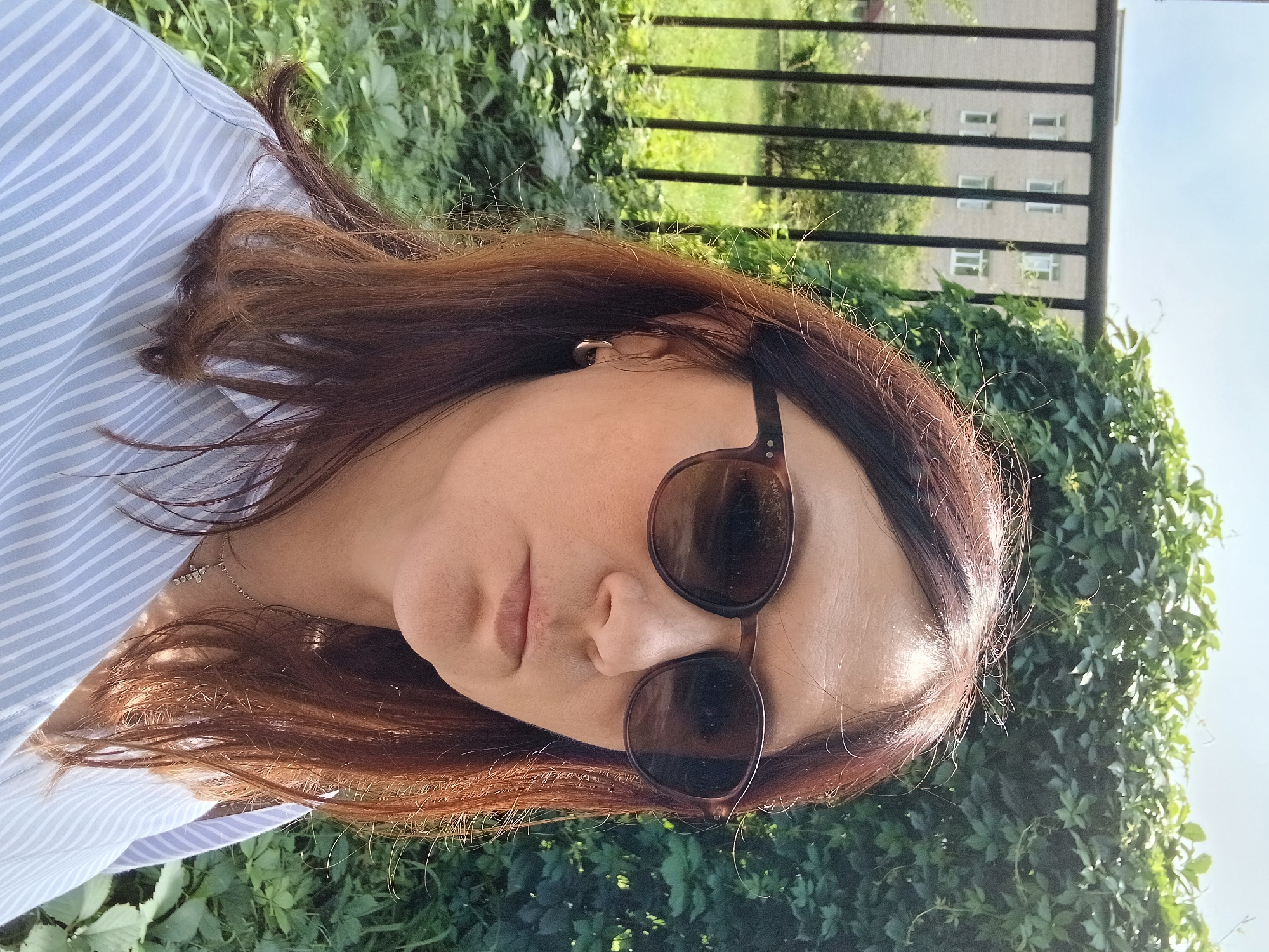 A medium shot captured on a bright, sunny day shows a woman with auburn hair, wearing large, round, brown-rimmed sunglasses and a light blue and white striped button-down shirt. She has a simple silver cross necklace with small gems visible around her neck and a small silver hoop earring in her visible ear. The woman is looking forward, her mouth slightly pursed. The background is lush green foliage, with a tall black metal fence behind it. Beyond the fence, a light-colored building with multiple windows can be seen, as well as some green grass and trees. The scene appears to be outdoors in an urban or suburban setting, possibly a park or residential area.