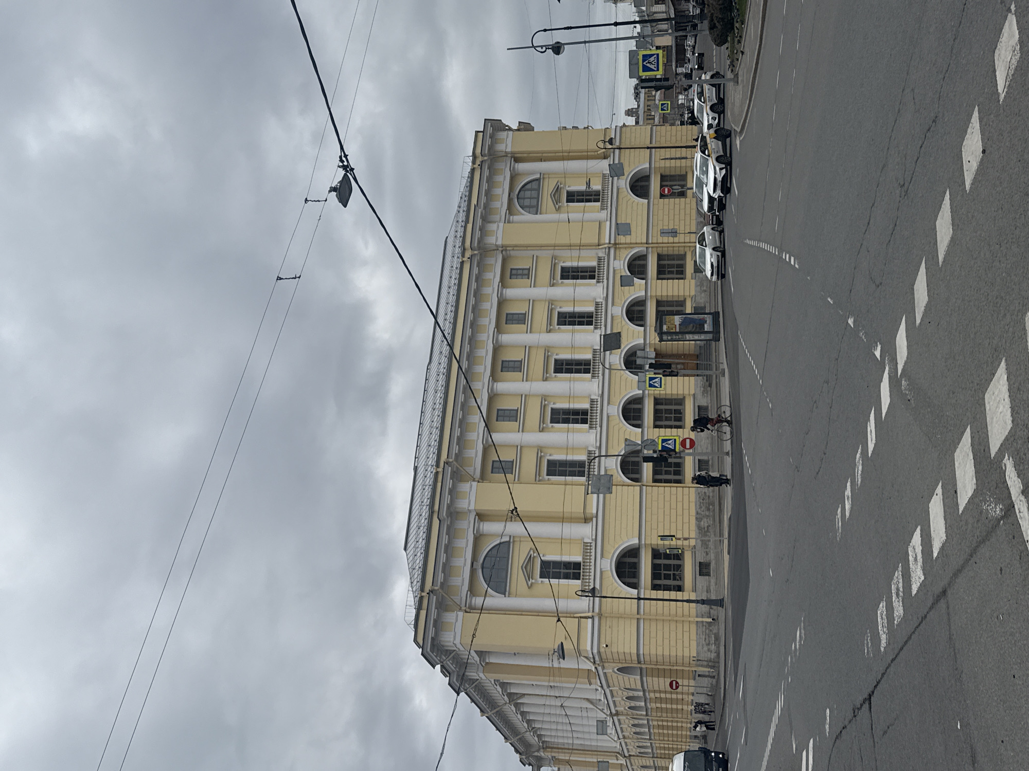 This image presents a street scene in Saint Petersburg, Russia, captured on an overcast day. The focal point is a grand, multi-story building on the left, adorned with a pale yellow facade and white classical pilasters. It features numerous windows, some rectangular and others with arched tops, and a glass-covered structure on its roof.
In the foreground, an asphalt road stretches into the distance, marked with white dashed lines and prominent white rectangular blocks, likely indicating a pedestrian crossing. Several white cars, including some identifiable as taxis by their yellow markings, are parked along the curb adjacent to the building. Overhead, a complex network of black wires crisscrosses the grey, cloudy sky, with a streetlamp fixture suspended among them.
Faint figures of people can be seen on the sidewalk and a cyclist on the road, engaging in routine urban activity. Street furniture includes a classic streetlamp post and various traffic signs: multiple blue square signs with a white pedestrian symbol (pedestrian crossing) and a red circular sign indicating 'No Entry'.
The time of day appears to be daytime, but the heavy cloud cover casts a muted light over the scene, contributing to a calm, everyday urban atmosphere. There is no specific event taking place, and no clearly legible text is visible on any signs or advertisements due to distance or blur.