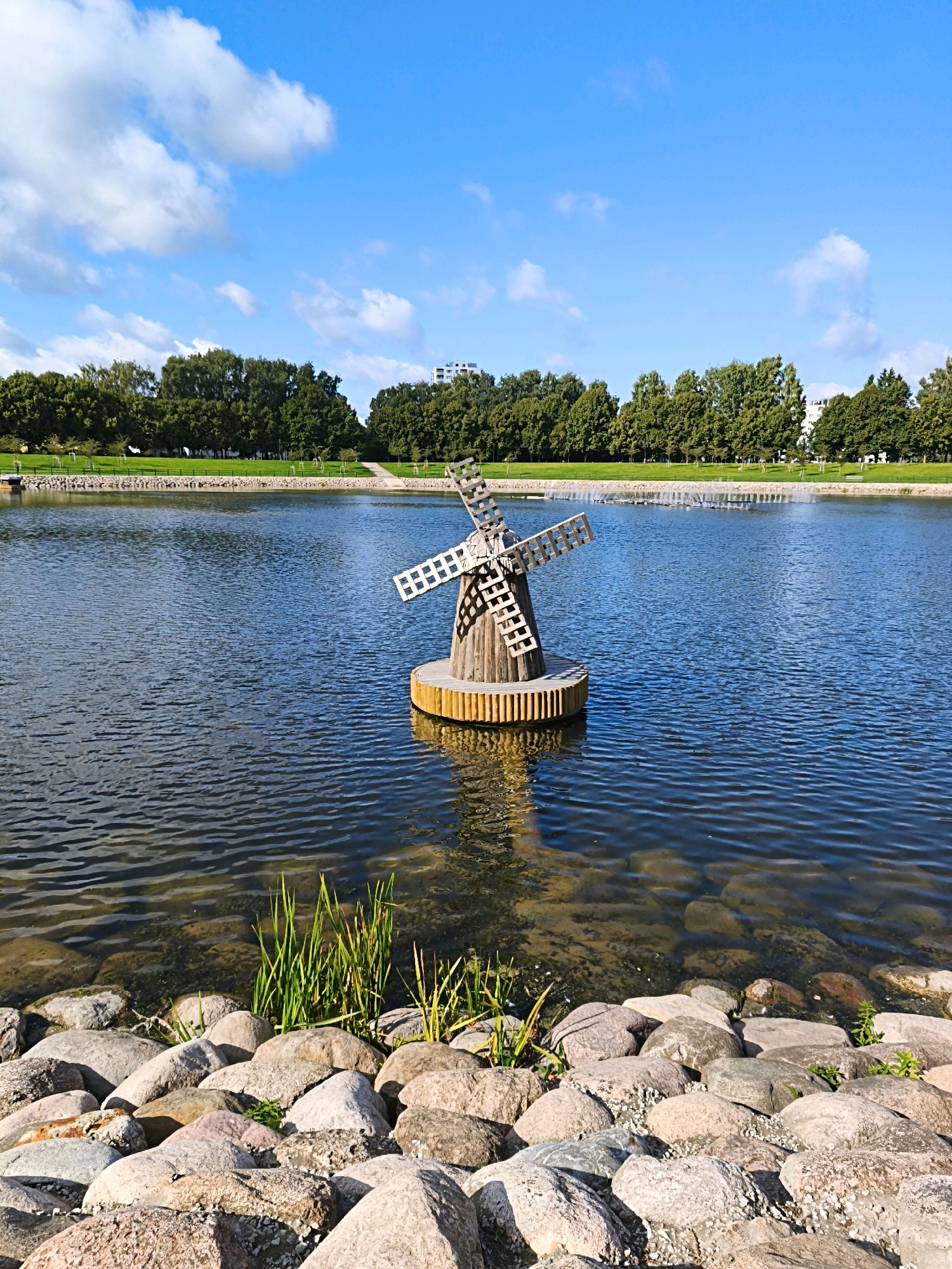 The image depicts a serene outdoor scene in Tallinn, Estonia, likely on a sunny day given the bright blue sky and visible clouds. In the foreground, a rocky shoreline lines a calm body of water, possibly a pond or lake. Among the rocks, some green reeds grow, adding a touch of natural texture.
Dominating the middle ground is a charming, small wooden windmill situated in the water. The windmill appears to be a decorative element, not a functional one, with its blades crafted from wood. It rests on a wooden platform that is partially submerged.
The background reveals a well-maintained park area with lush green grass and a line of mature trees. Beyond the trees, modern buildings can be seen, suggesting an urban setting. In the water, further in the background, a series of fountains are active, creating a gentle spray. The overall atmosphere is peaceful and picturesque, characteristic of a public park or waterfront area in the city. There are no people or animals visible in the frame.