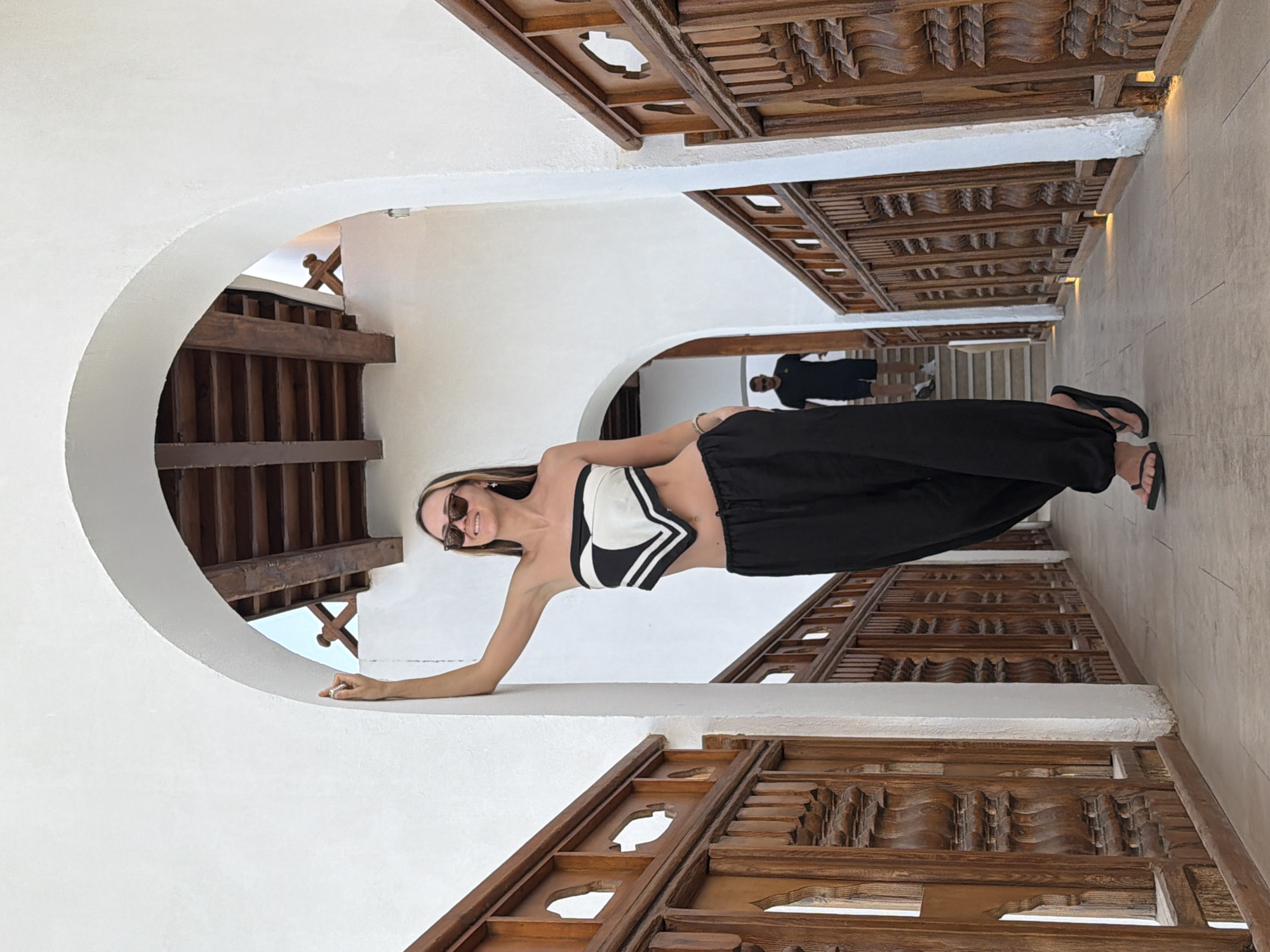 The photograph captures a woman in a black and white striped top and black pants, leaning against the wall of a structure with a circular archway. She is wearing sunglasses and sandals, and there is a man in the background. The setting appears to be a corridor or walkway, characterized by detailed wooden architectural elements, possibly of Islamic design. The location can be identified as Sharm El Sheikh, Egypt, given the style of architecture. The building seems to be a hotel or resort.