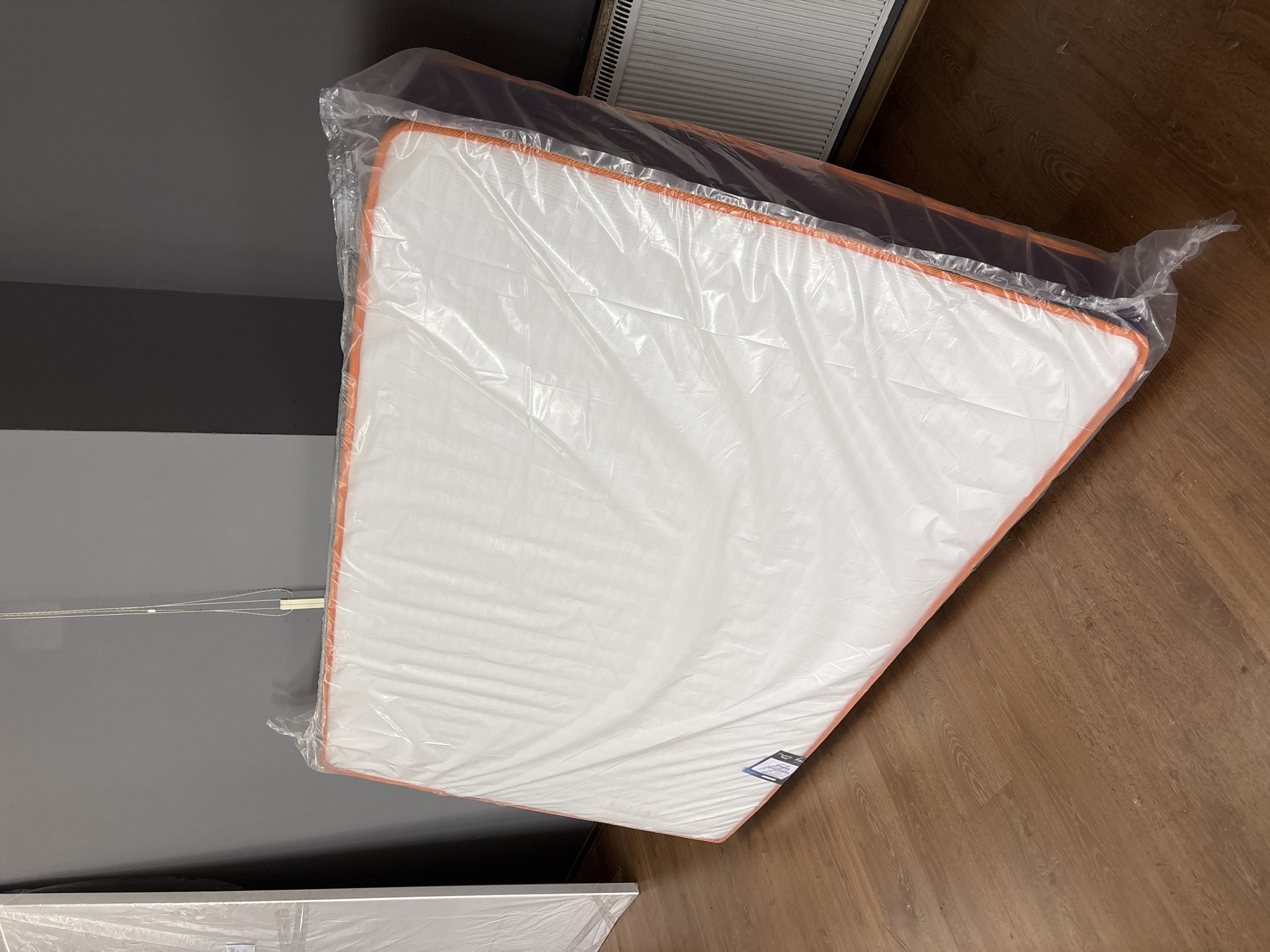 A mattress, wrapped in clear plastic, is propped up against a dark gray wall. The mattress has an orange trim around its edges. It is lying on a brown wooden floor. To the left of the mattress, another white object, also wrapped in plastic, is visible. In the background, a white radiator with vertical slats is present. The scene is indoors, likely in a room or storage area.