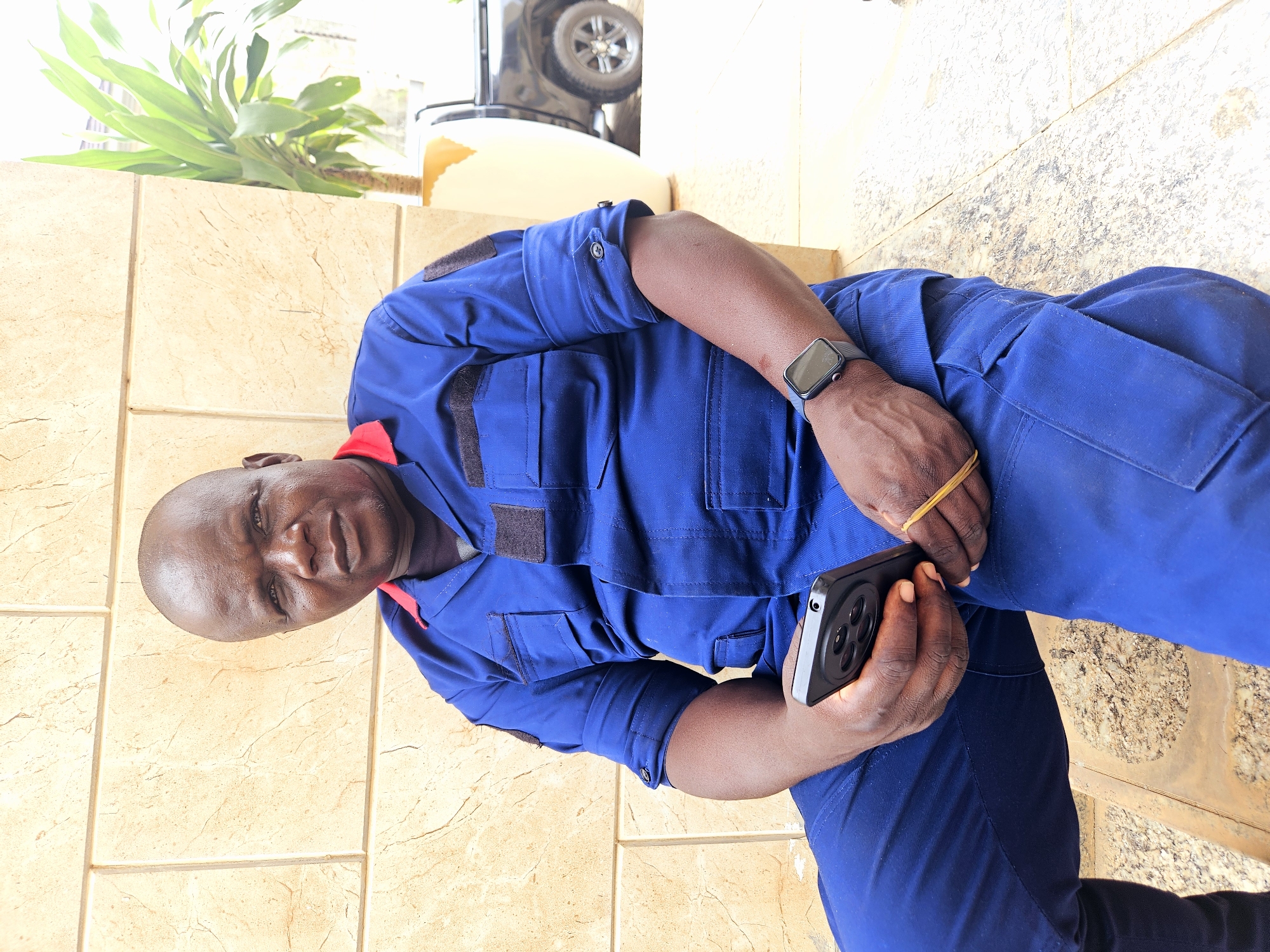 A medium shot captures a bald, dark-skinned Black man in a blue uniform with red trim, seated outdoors and looking directly at the camera. He is wearing a dark undershirt and the uniform has multiple pockets with dark patches. On his left wrist, he wears a dark smartwatch with a square face. He holds a black smartphone with a circular camera array in his hands, which are resting on his lap. A yellow rubber band is visible on his right wrist. The man is seated on a concrete structure in front of a light brown tiled wall. To his right, a large potted plant with green leaves is visible, and behind it, a black truck is partially visible. The overall setting suggests an outdoor location in the city of Guzape, Nigeria.