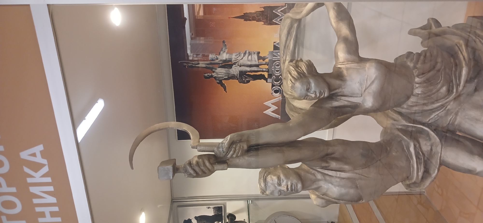 The image shows a display case containing a replica of the iconic Soviet statue "Worker and Kolkhoz Woman." The statue features two figures: a man (the worker) holding a hammer, and a woman (the kolkhoz woman) holding a sickle. Together, they hold the hammer and sickle aloft. In the background behind the statue replica, there is a photograph or print depicting the original statue with a cityscape, presumably Moscow, in the background. The photograph includes text that says 'Moscow' in Russian. The display case appears to be in a museum or exhibition hall, given the presence of informational signage and other displays visible. The statue is made of metal.