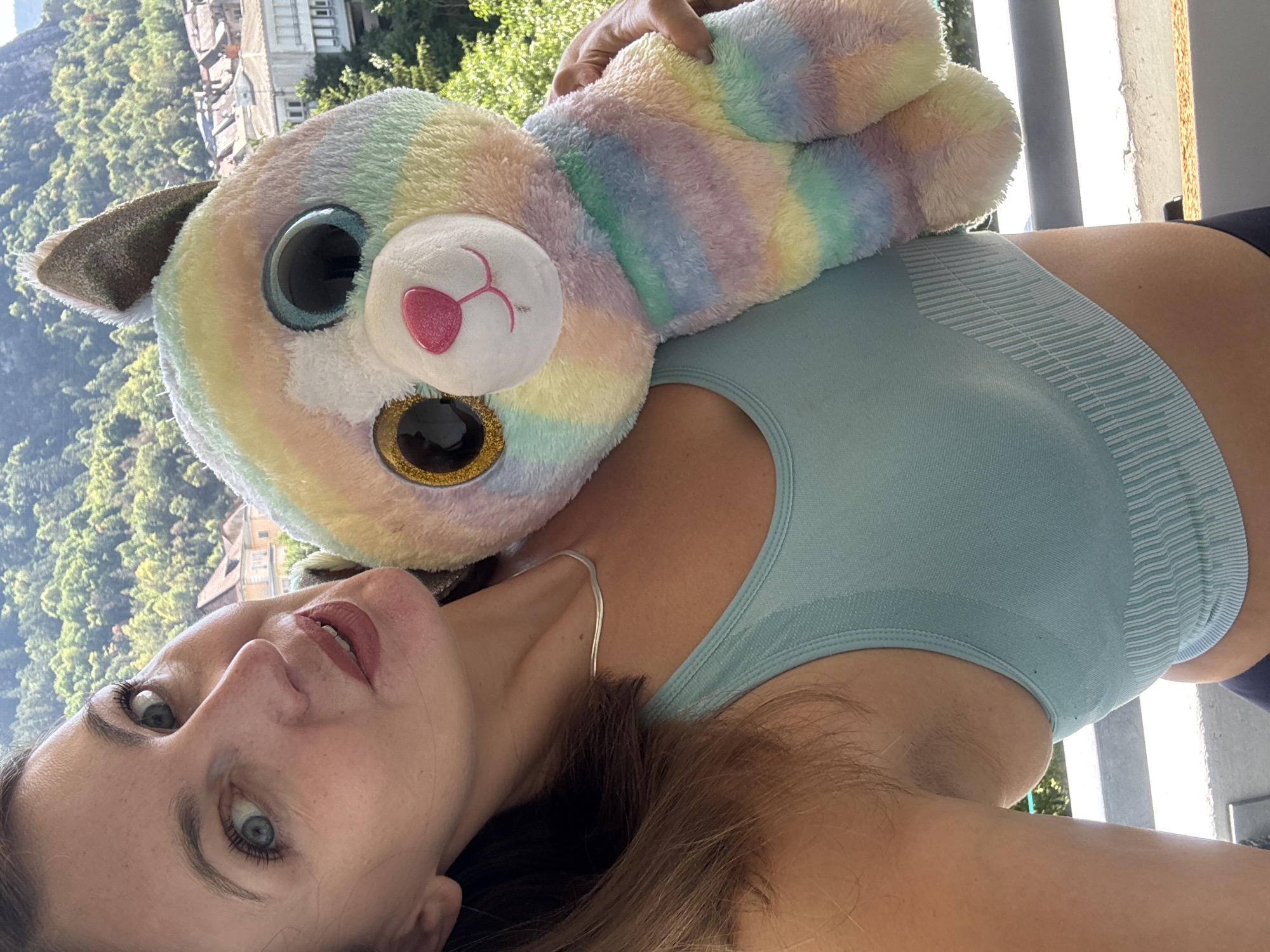 A woman is holding a rainbow-colored plush cat toy on a balcony in Saint-Claude, France. She is wearing a light blue sports bra and a thin silver necklace. The plush toy has large, glittery eyes and a pink nose. The background shows a hillside with buildings and lush green trees. The woman is looking at the camera with her head tilted. The lighting suggests it is daytime.