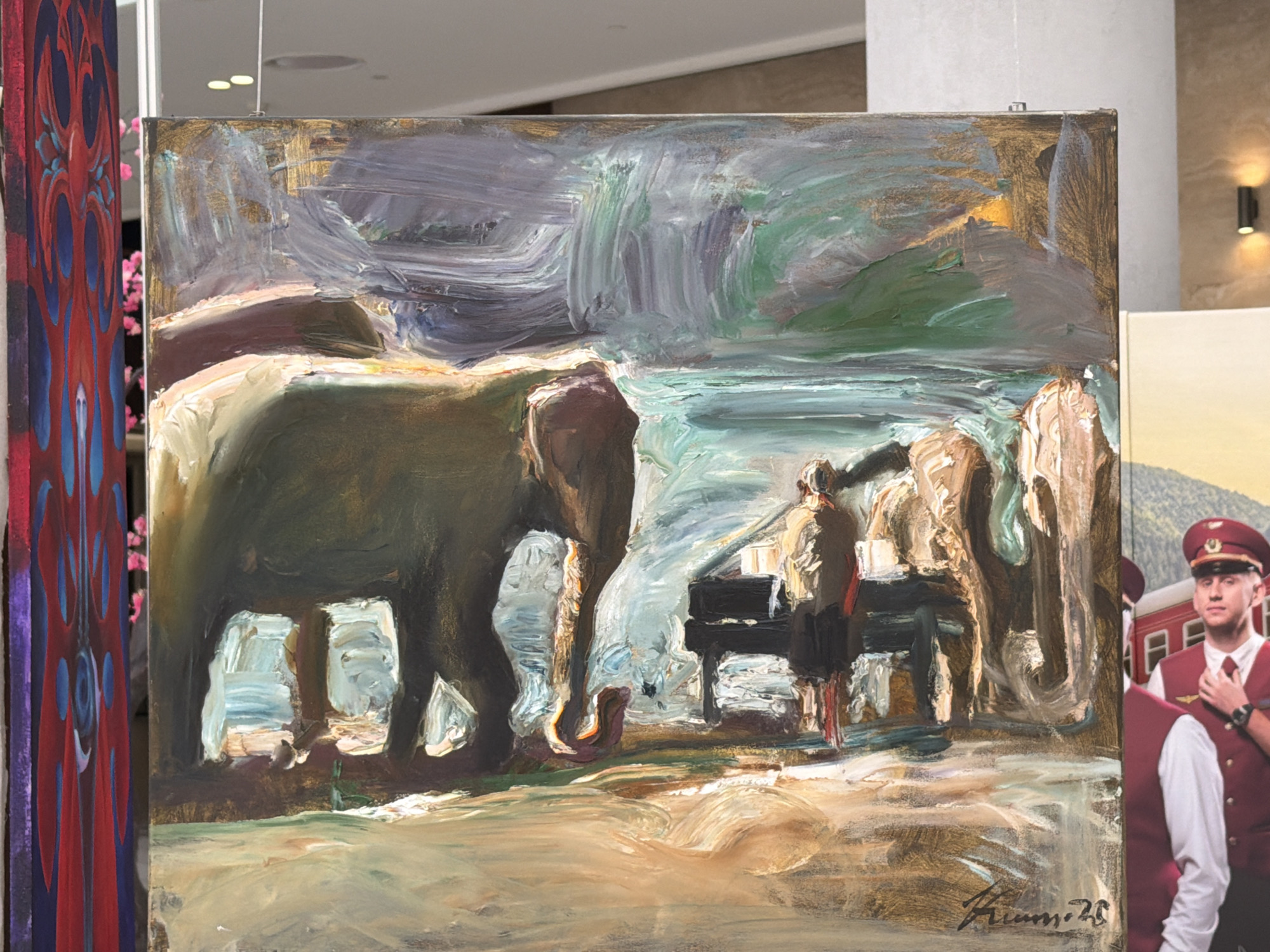 The image shows a painting of elephants playing a piano in what appears to be a gallery or exhibition space. The painting features a large elephant on the left, with two smaller elephants to the right, all gathered around a grand piano. Two figures, possibly people, are also present near the piano. The background is abstract, with swirls of color and texture suggesting a landscape or atmospheric effect. The painting is signed in the lower right corner with a signature that appears to read "Himmi 23". To the left of the painting is another artwork with a red and blue abstract design. To the right of the painting, a section of another artwork shows a man in a red uniform and cap, possibly a train conductor, standing in front of a red train. The scene is illuminated by artificial lighting. The context provided indicates this scene is located in the city of Kyiv, Ukraine.