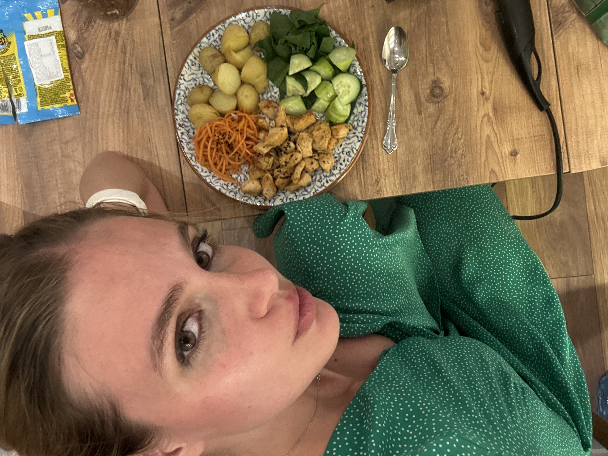 A person wearing a green, polka-dotted dress is lying on a wooden floor next to a plate of food. The plate contains boiled potatoes, sliced cucumbers, cooked shredded carrots, sauteed chicken pieces, and spinach. A spoon sits to the right of the plate. A bag of snacks is visible in the upper left corner of the frame. A dark, curved object with a cord, possibly a hair dryer, is in the upper right corner. The location is Unknown, Unknown.