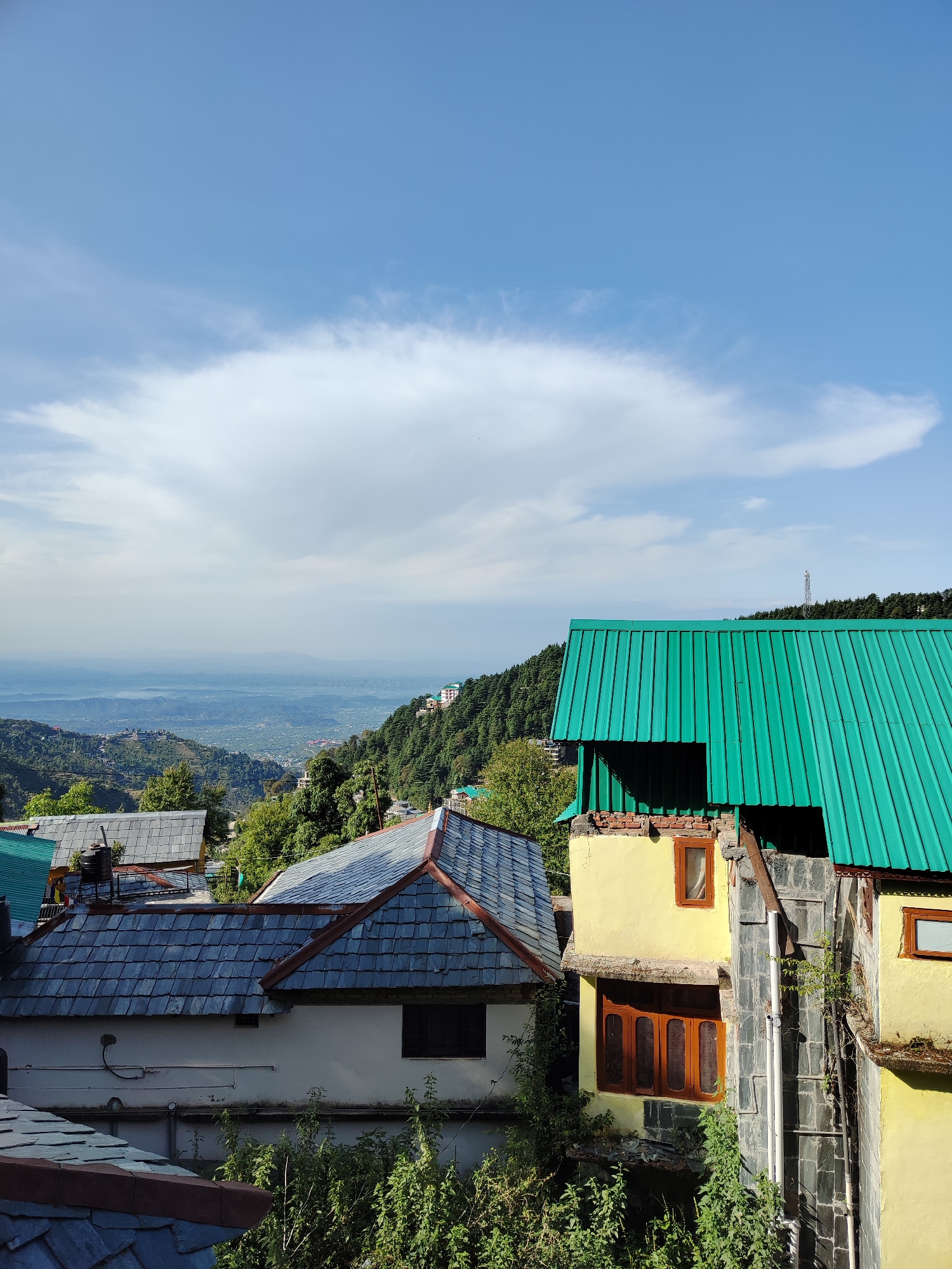 The image depicts a hillside view of buildings with various colored roofs in what appears to be a mountainous region, possibly in the city of Haini, India. The foreground includes a yellow building with a green corrugated metal roof and a stone-tiled roofed building. The background shows distant mountains shrouded in haze and a blue sky with clouds. Lush green trees cover the slopes, with a mix of residential and commercial structures visible, indicative of a village or small town.